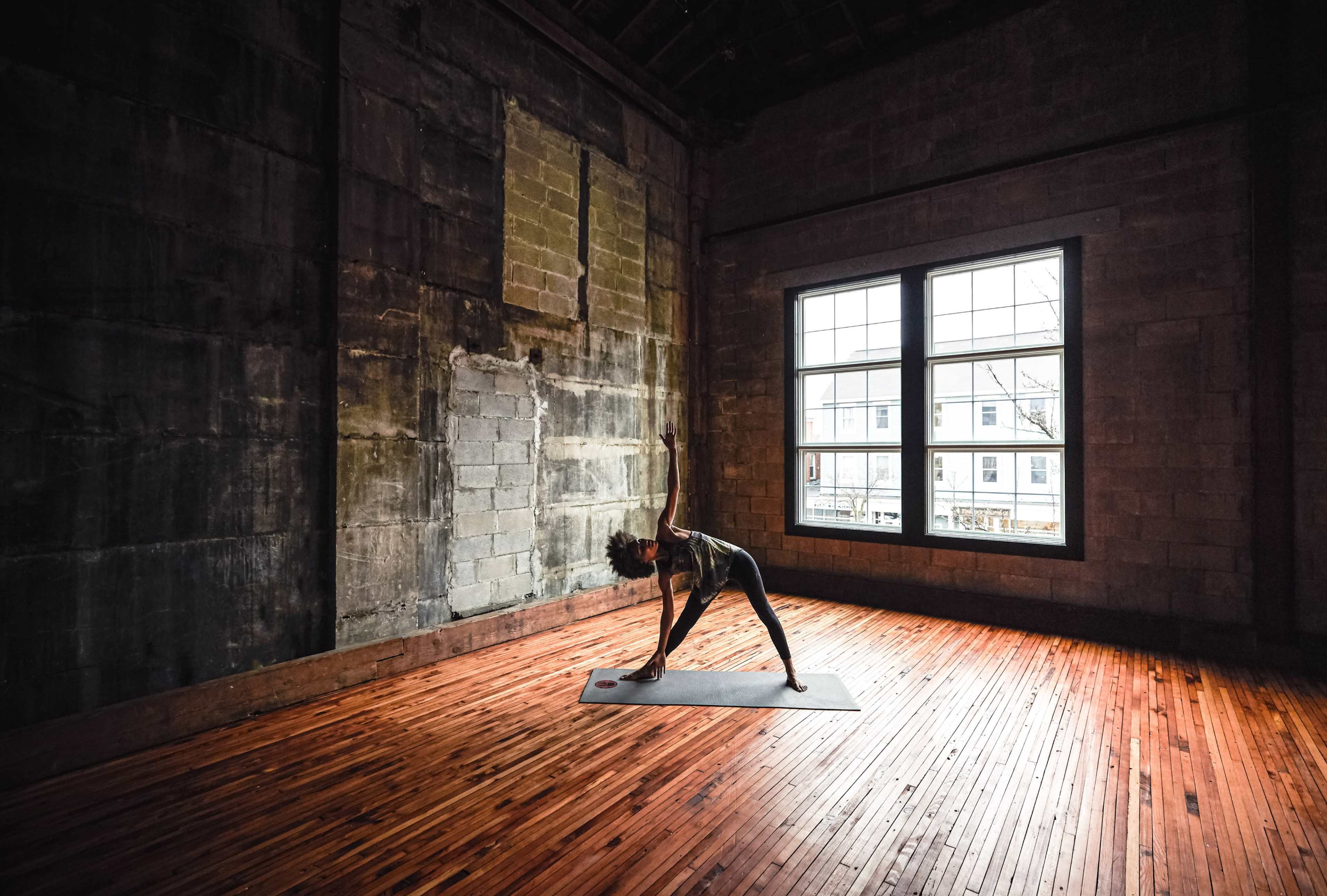 A person is performing a yoga pose on a mat in a room with large windows and wooden flooring.