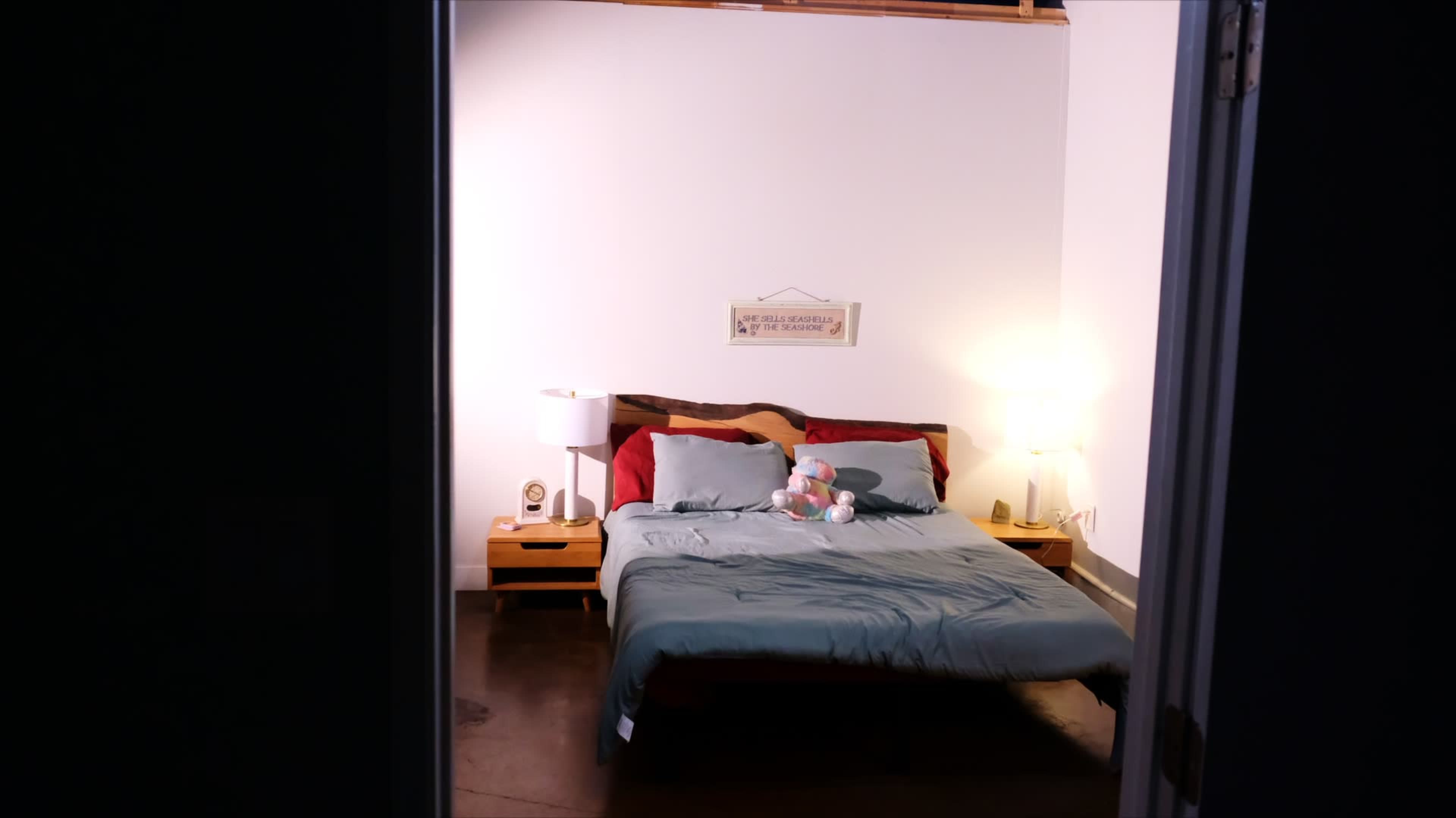 A neatly made bed with a blue comforter, plush toys, and bedside lamps is visible in a warmly lit bedroom.