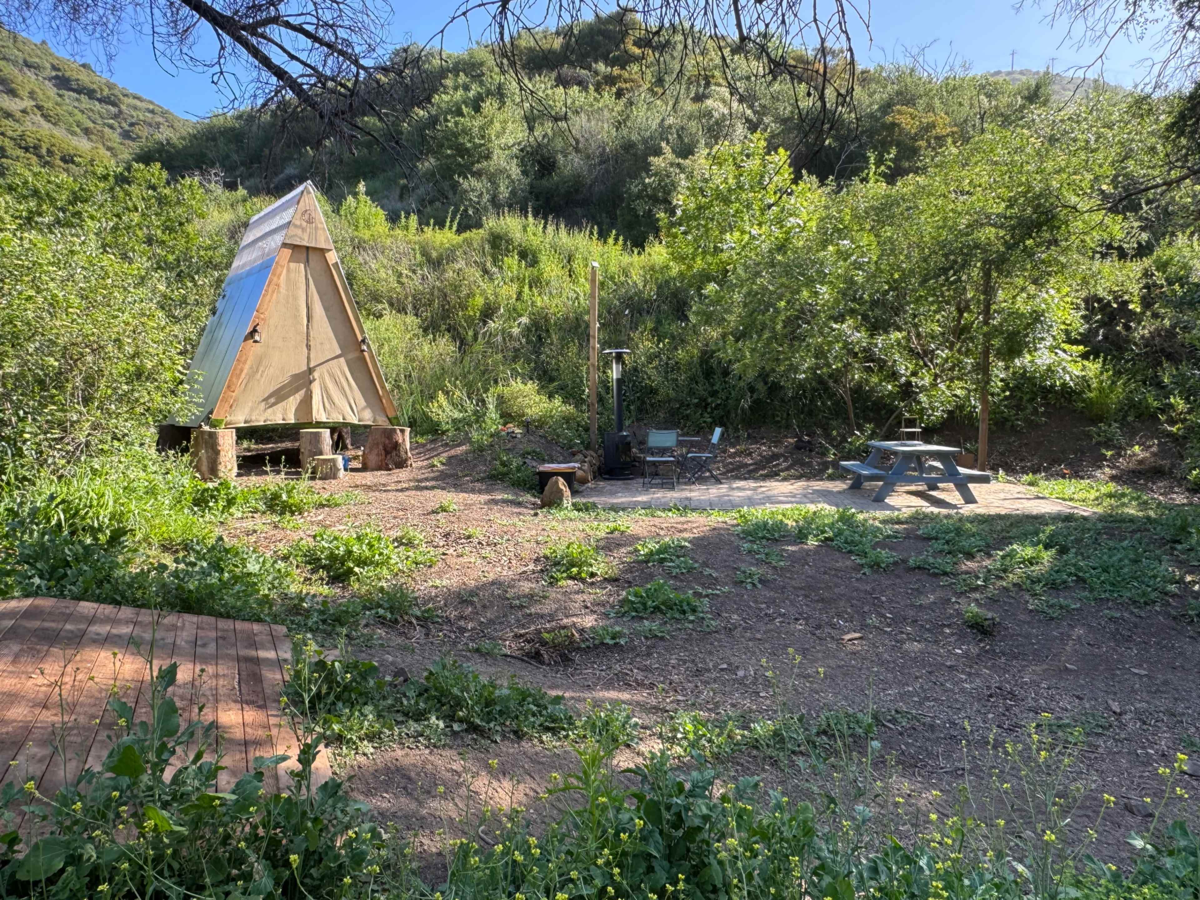 The image shows an open area with a triangular wooden structure, picnic table, and fire pit, surrounded by greenery and hills in the background.