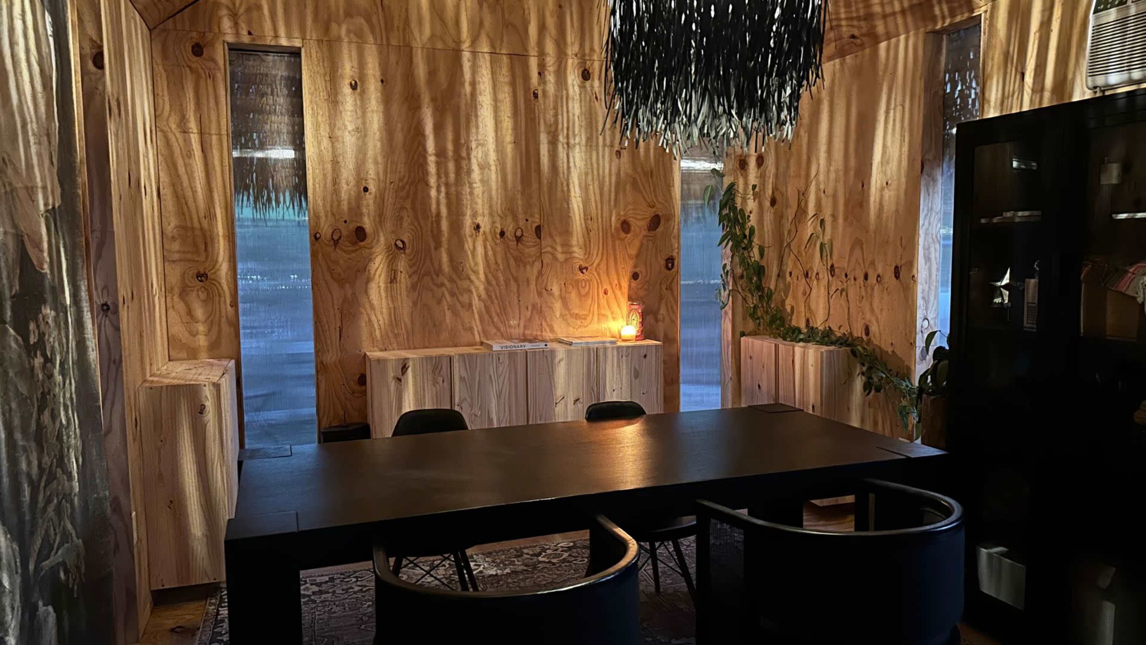The image depicts a dining room with wooden walls, a large black table surrounded by black chairs, and a unique chandelier overhead, featuring a candle on a side table.