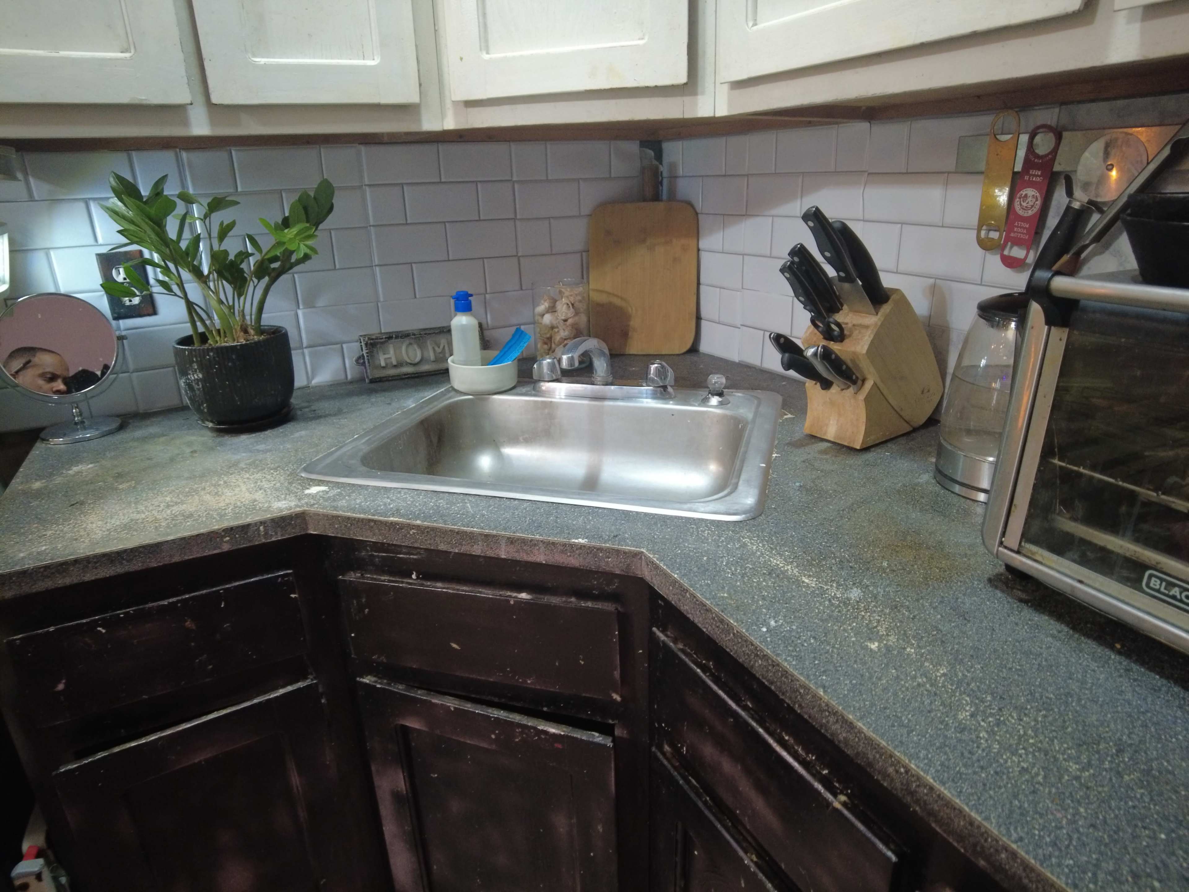 A corner kitchen area features a stainless steel sink, a plant, a knife block, and various kitchen appliances against a tiled backsplash.