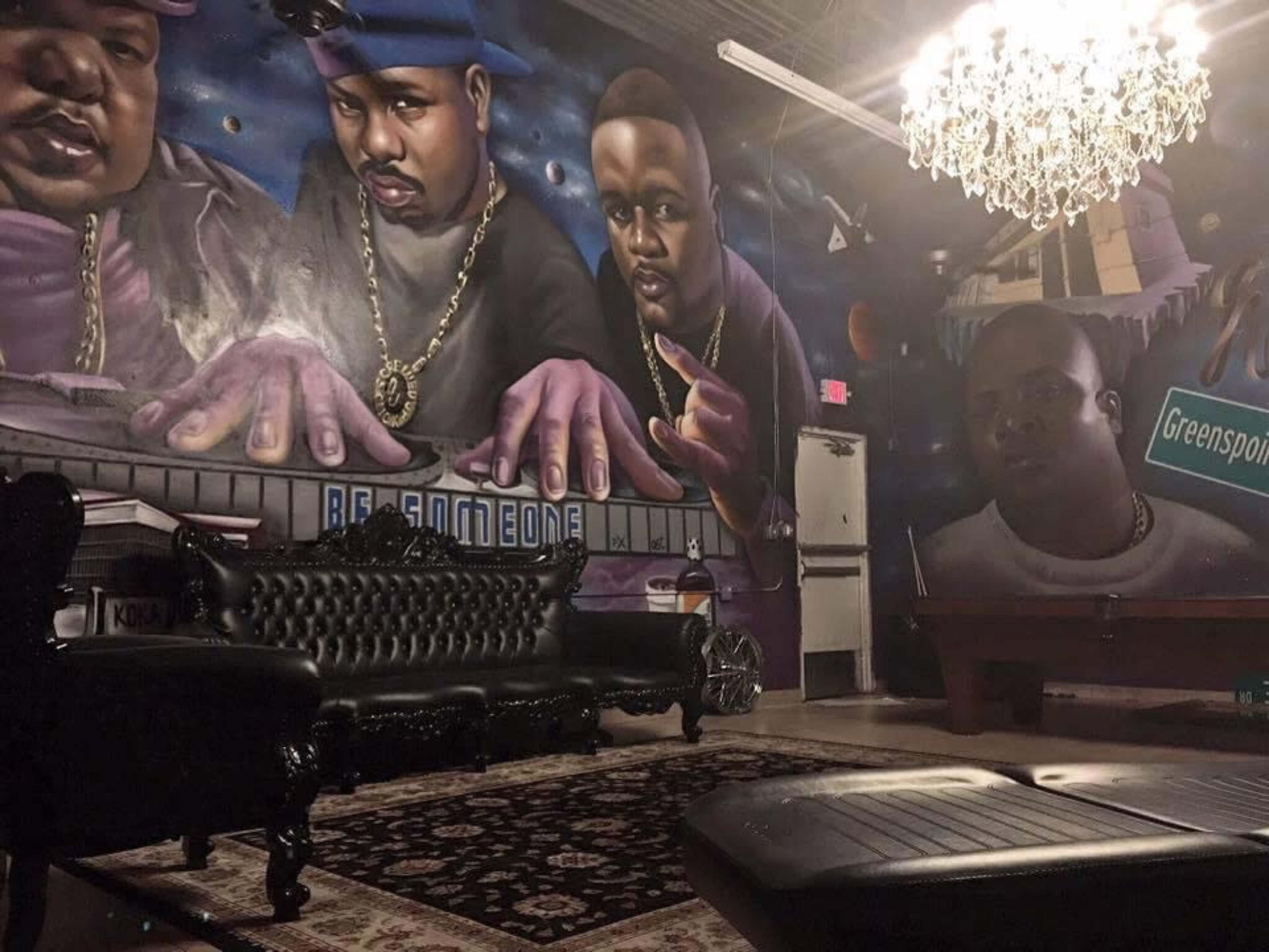A room with a mural depicting three men, with a black leather couch and a chandelier overhead, alongside a pool table and a patterned rug.