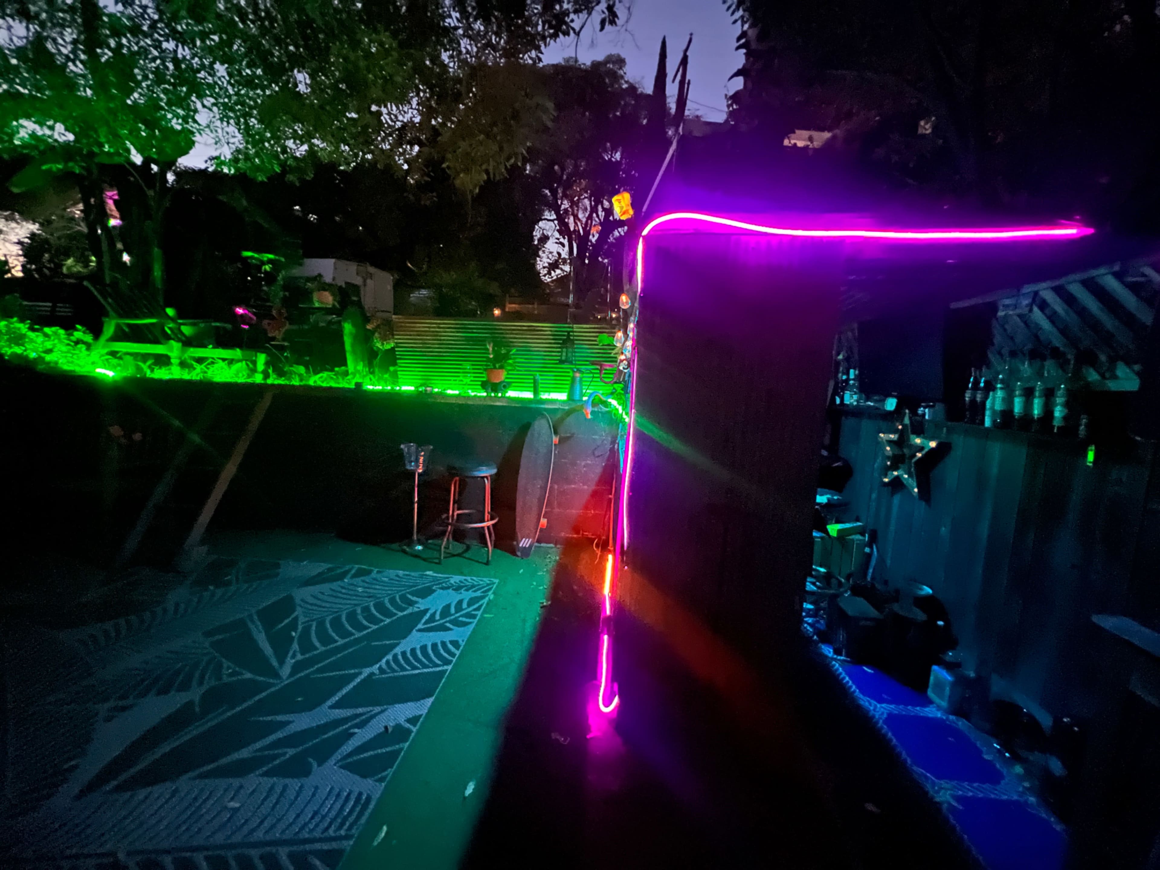 A dimly lit outdoor space with neon green and pink lights outlining structures and pathways among trees.