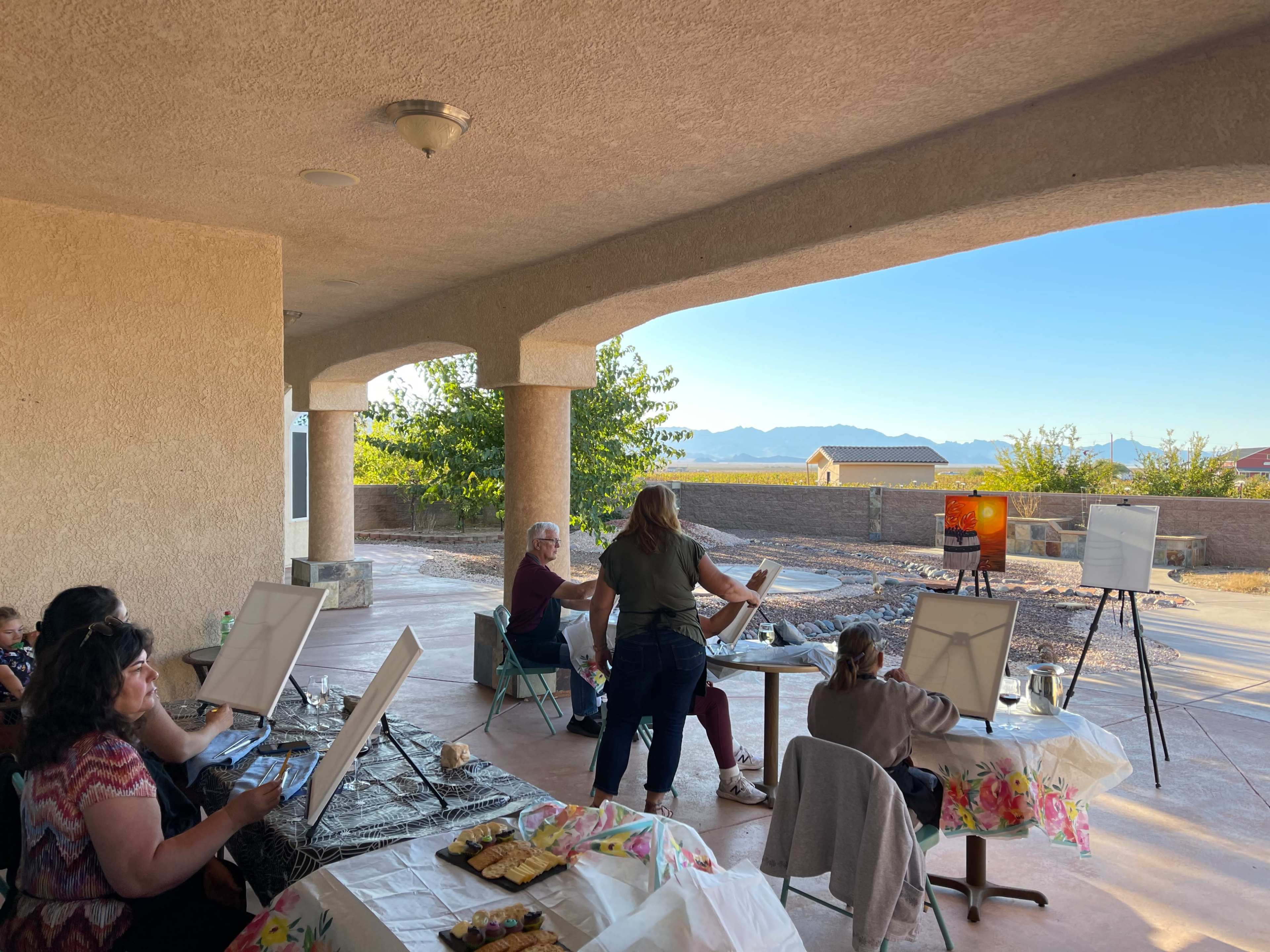 A group of people participates in a painting class outdoors under a covered patio with a mountain view.