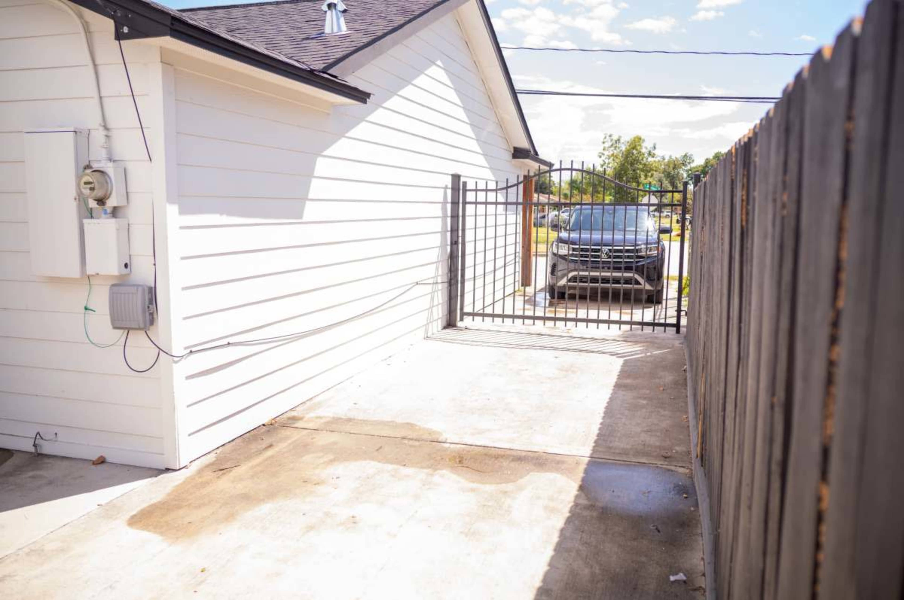 The image shows a concrete driveway leading to a gated entry beside a white house, with a vehicle visible in the background.