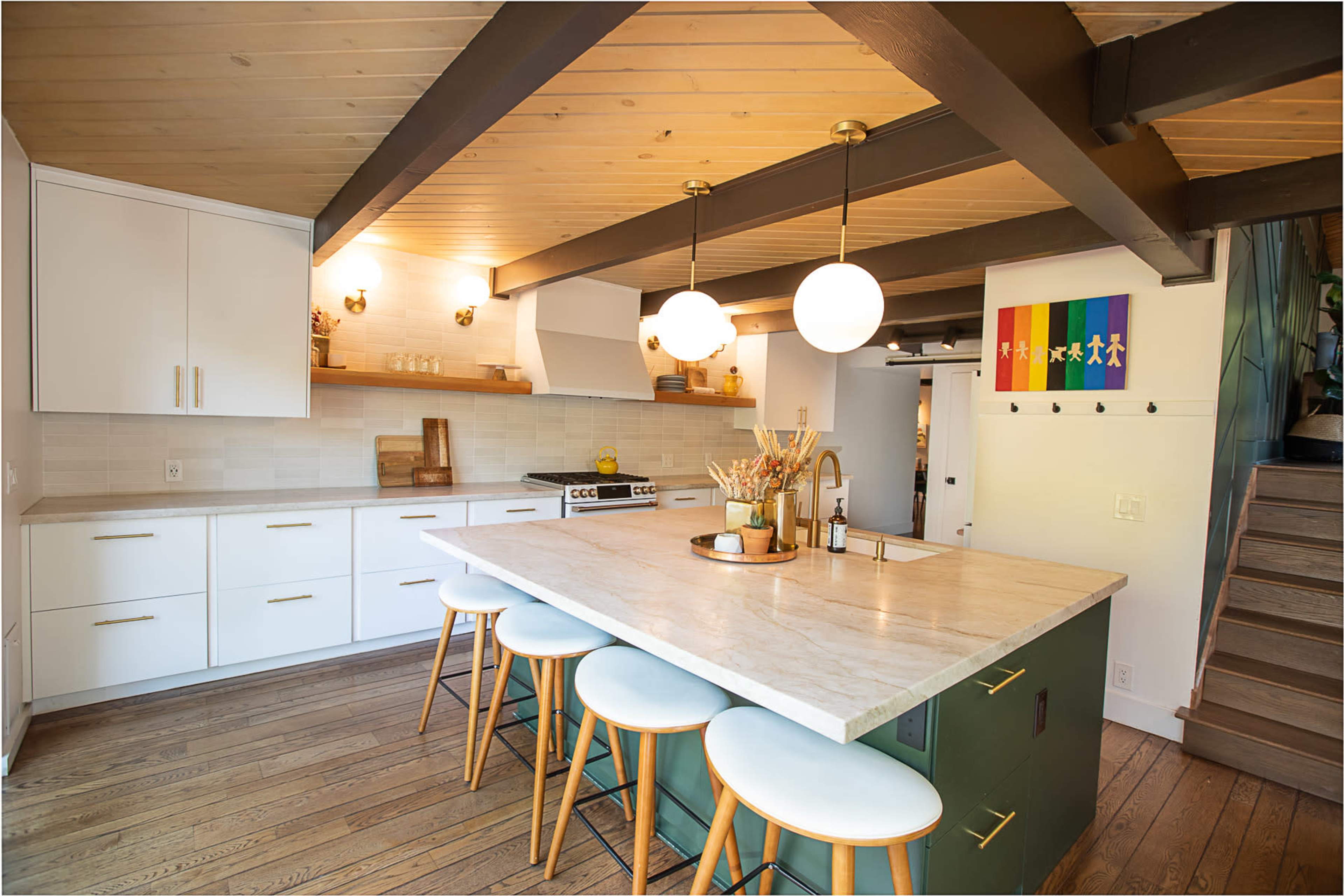 A modern kitchen features a central island with seating, white cabinetry, wooden beams, and a colorful wall decoration.