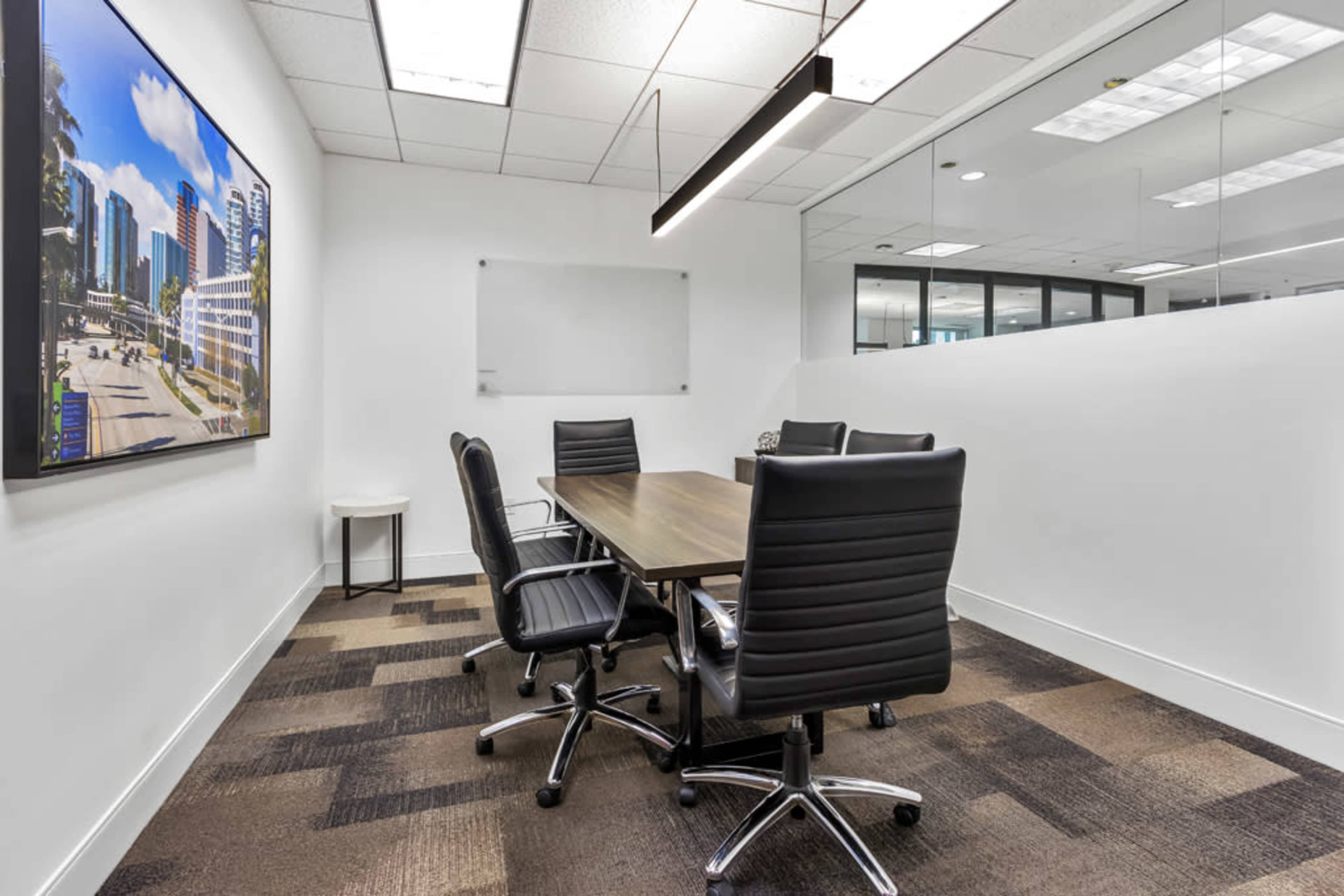 The image shows a modern conference room with a rectangular table and four black chairs, featuring a large wall-mounted screen displaying a cityscape.