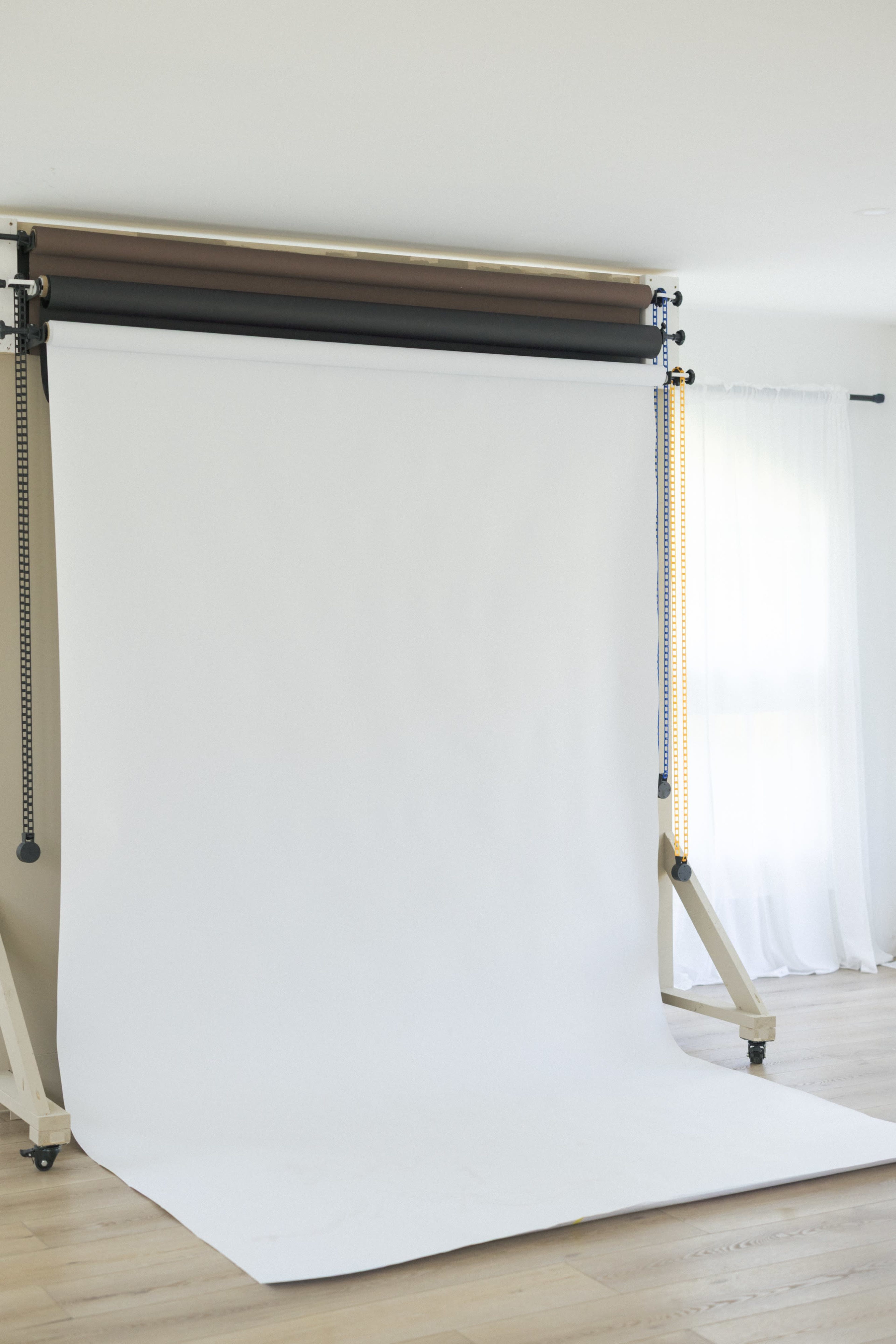 A blank white backdrop is set up on a stand in a well-lit room with wooden flooring and a sheer curtain.