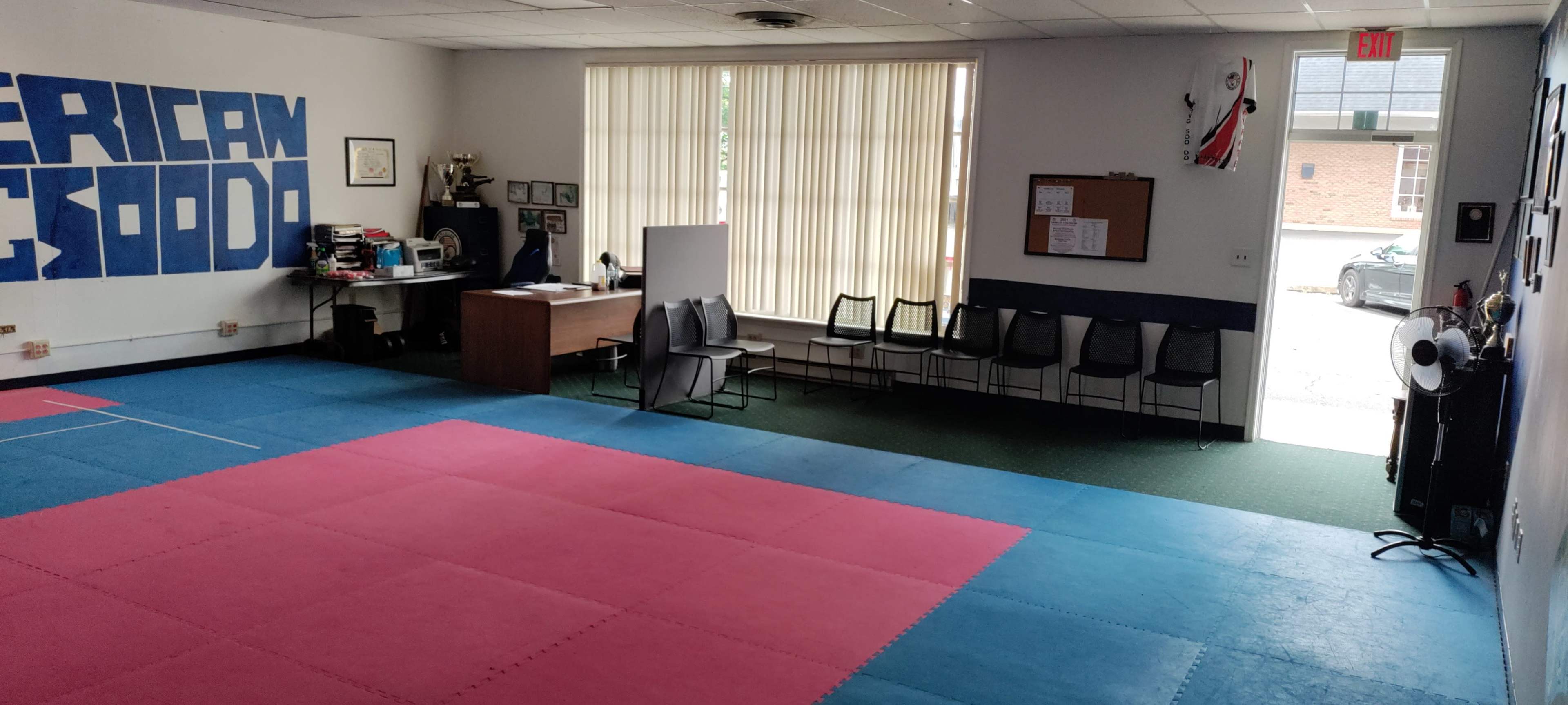 A sparsely furnished martial arts studio with blue and pink mats on the floor, a desk, and several chairs along the wall.