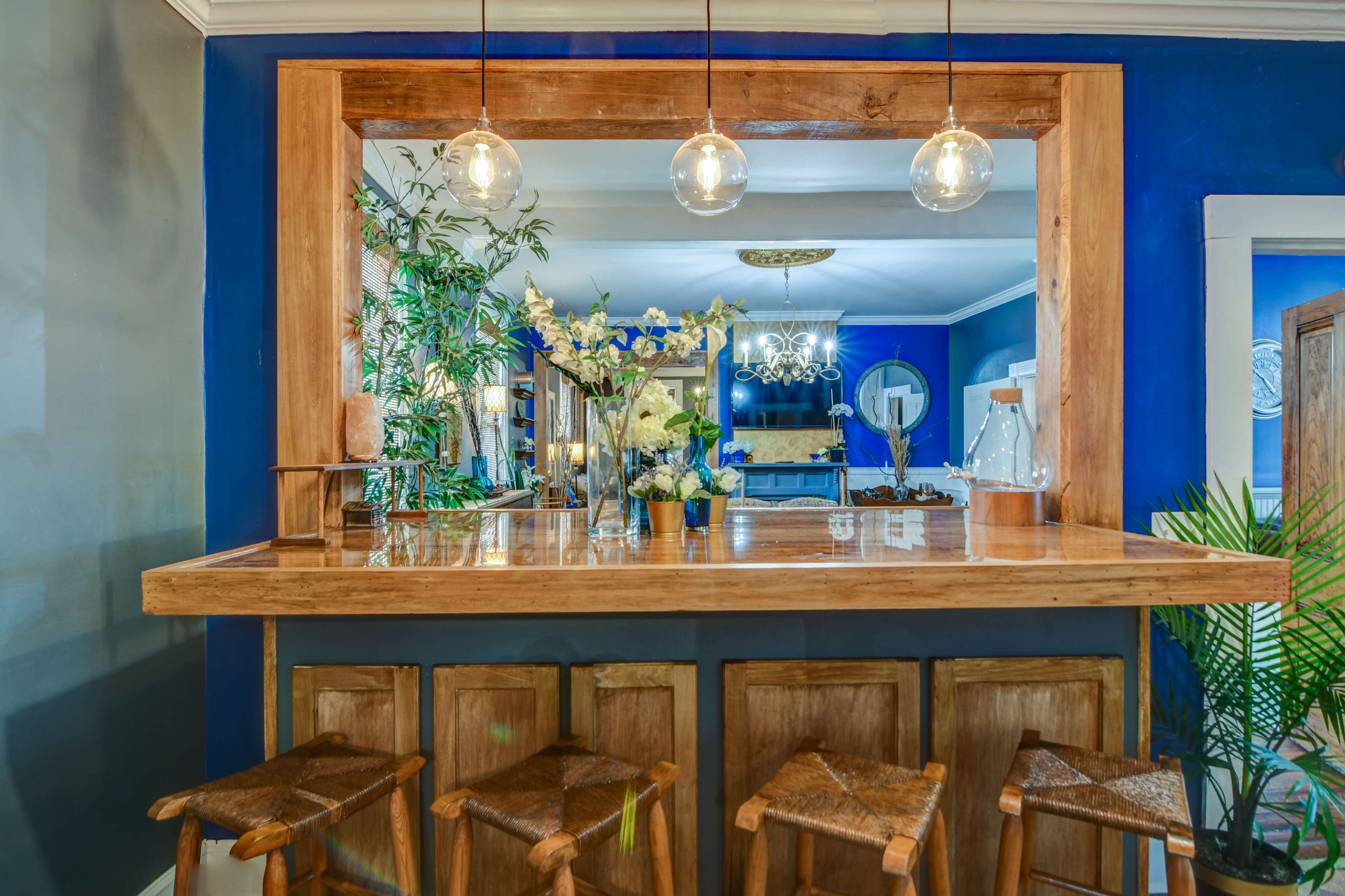 A wooden bar with three stools is lit by pendant lights and features a vase of flowers, set against a blue wall with plants in the background.