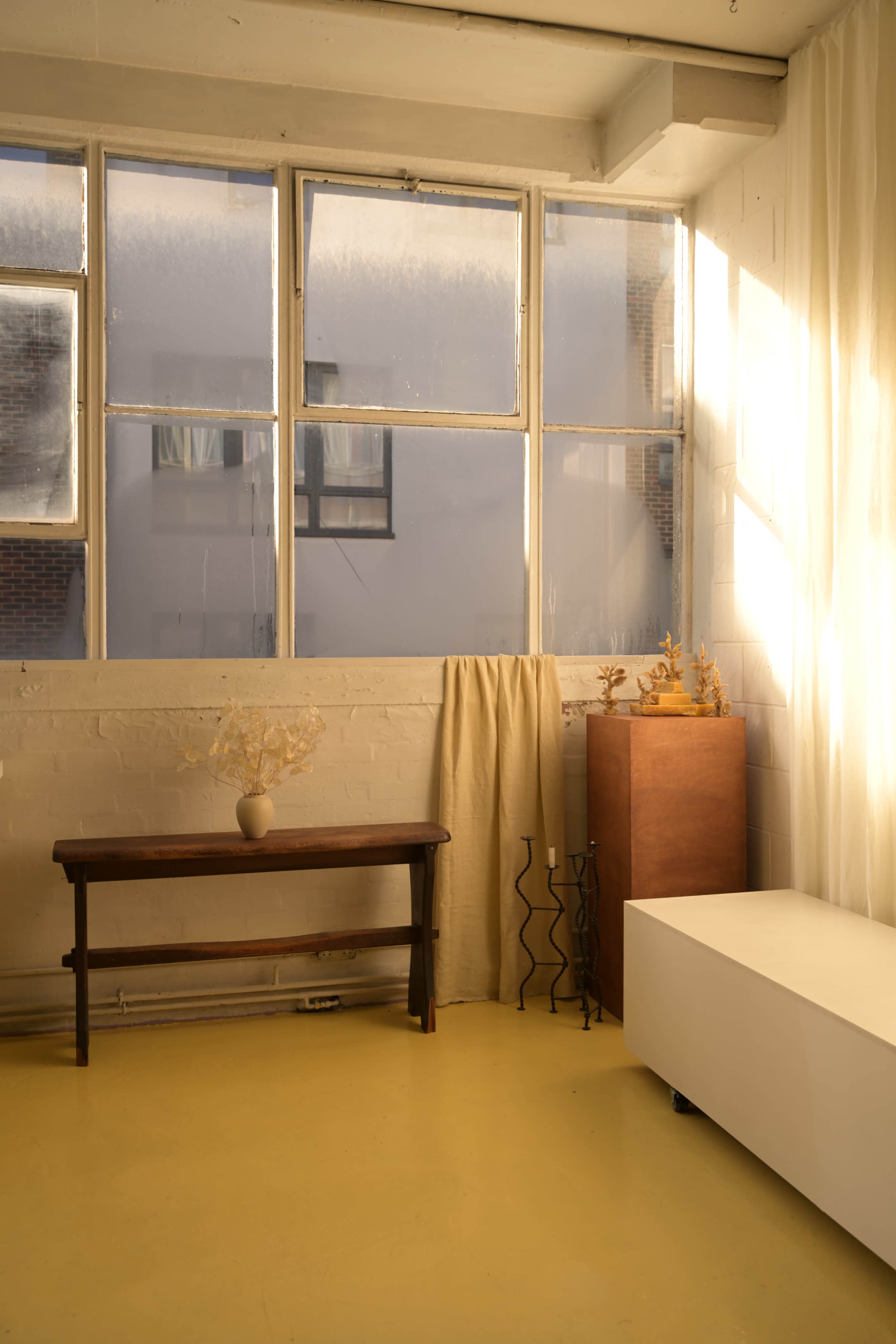 The image shows a bright room with large windows, a wooden table with a vase, a curtain, a wooden cabinet, and a white furniture piece on a yellow floor.