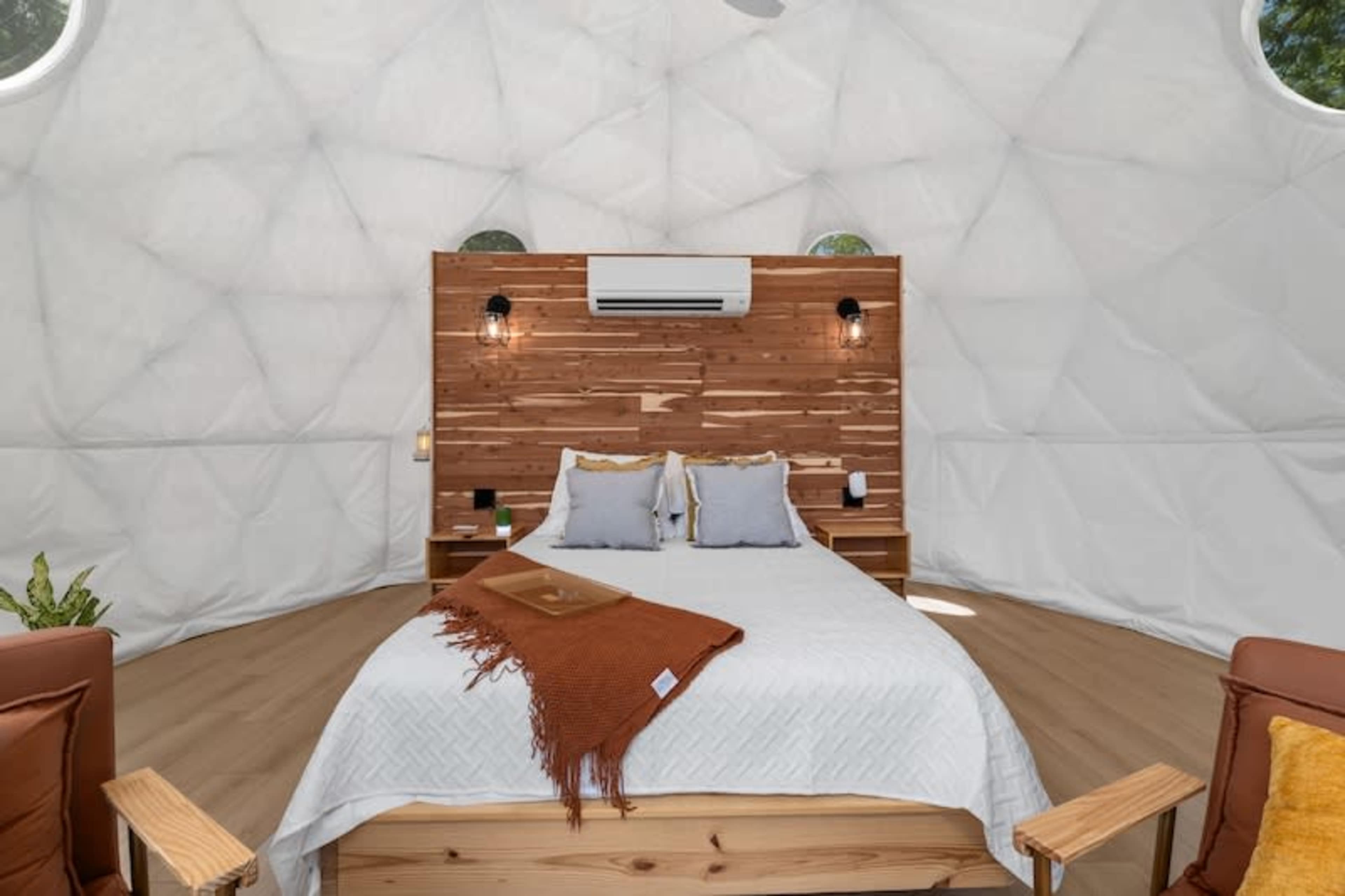 A geodesic dome interior features a wooden accent wall, a queen-sized bed with gray linens, and cozy seating areas.