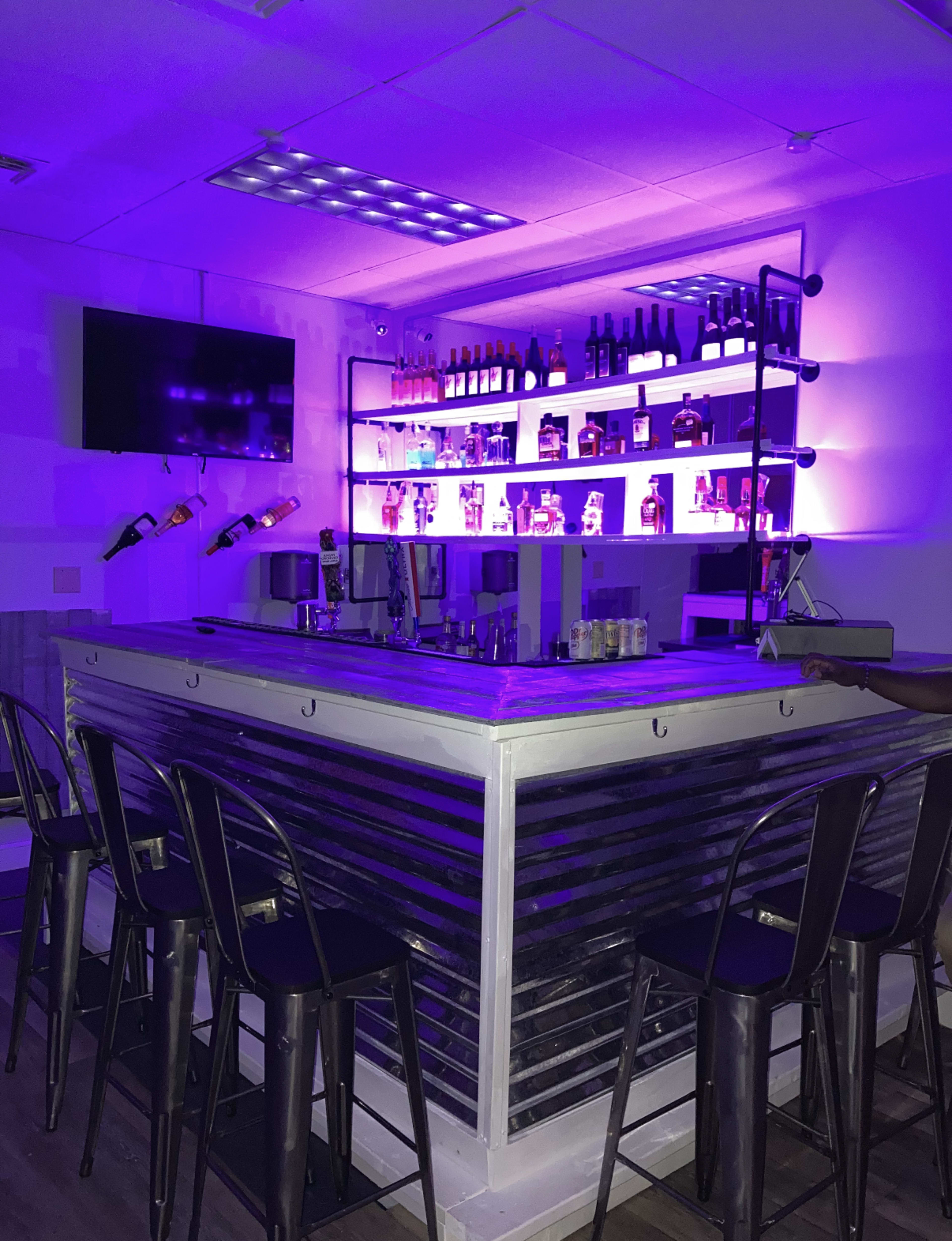 A modern bar with purple lighting, showcasing bottles of liquor on shelves behind a metal and wood counter, is surrounded by black metal stools.