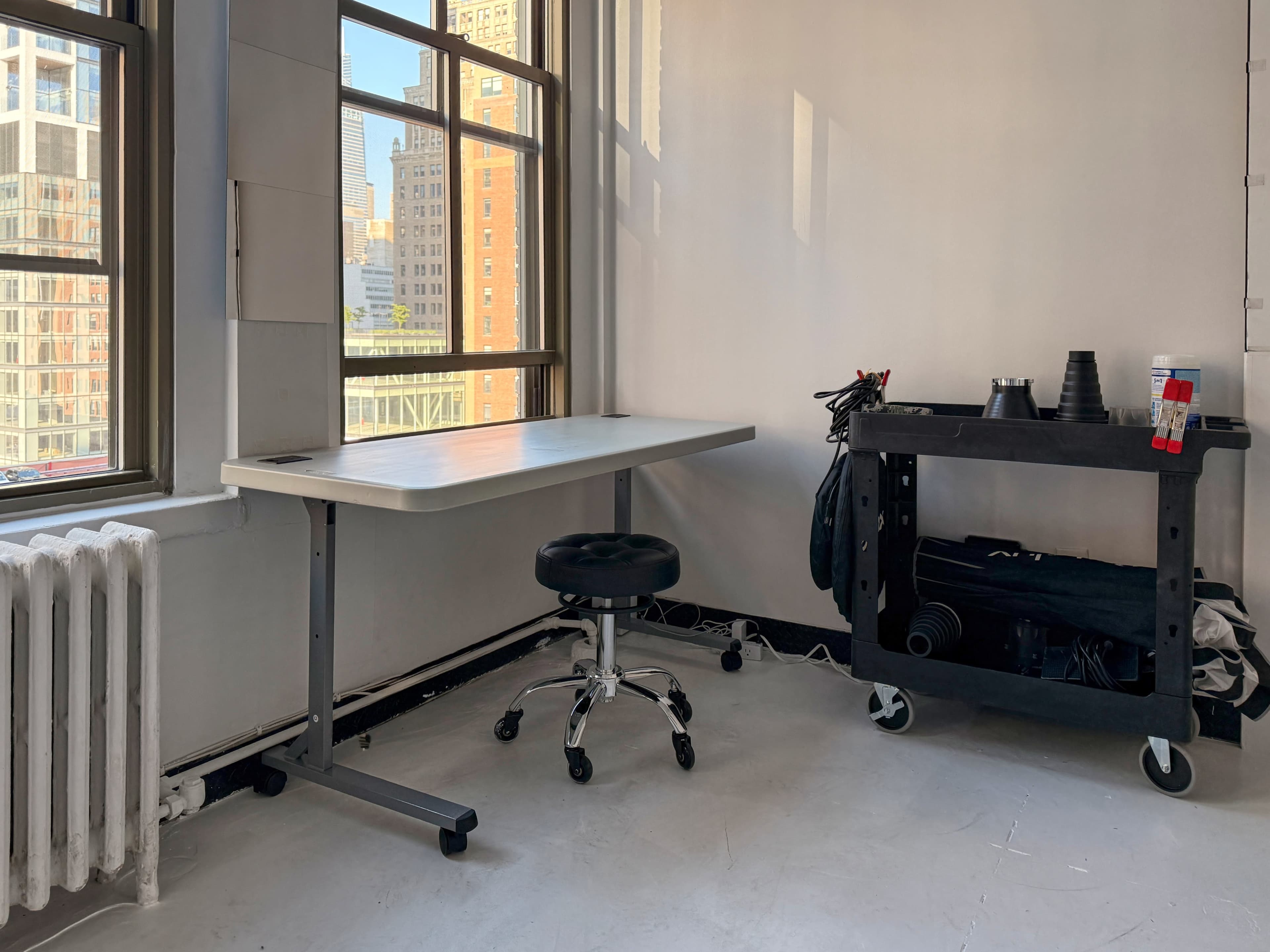 A simple workspace features a white table, a rolling black stool, and a black cart with various items, all set against large windows letting in natural light.