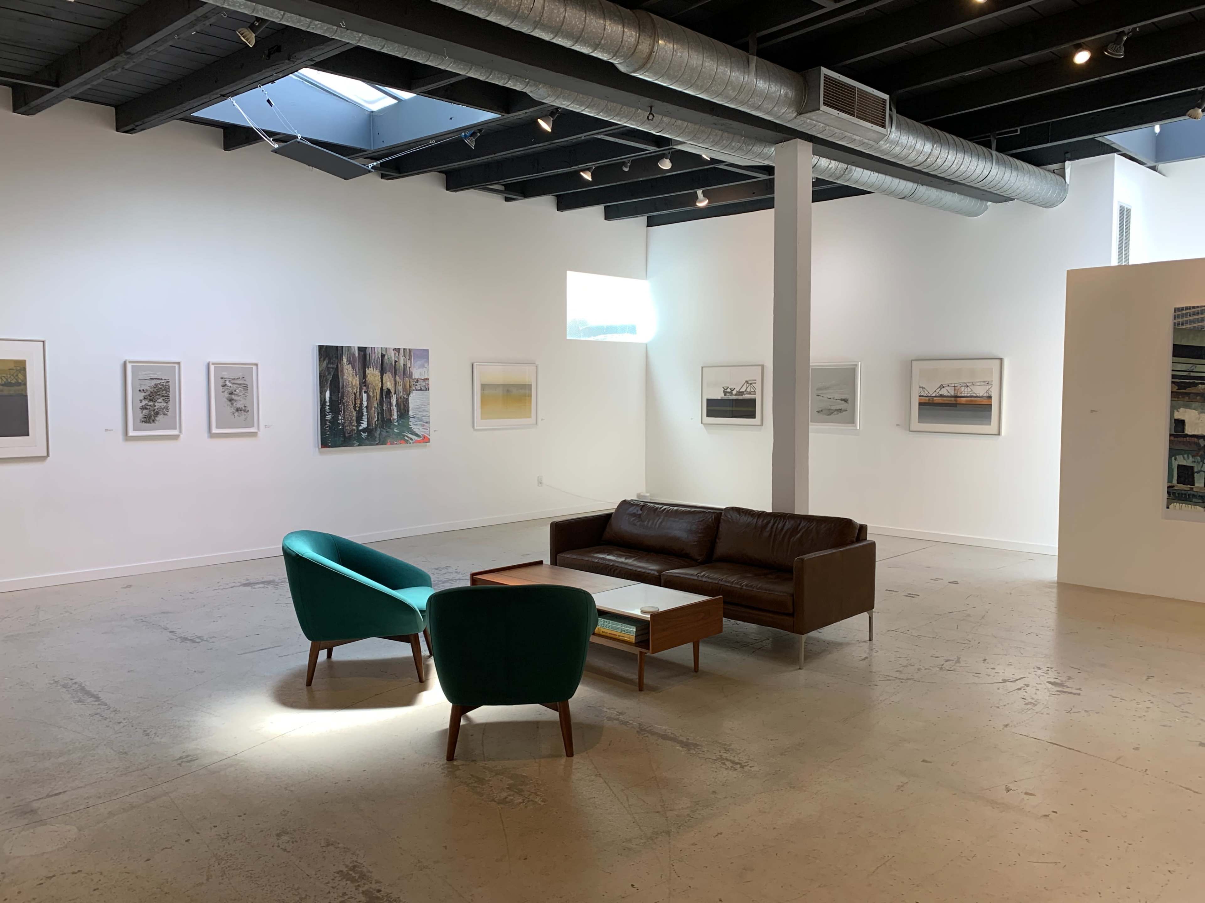 The image shows an art gallery featuring white walls adorned with framed artwork, a brown leather sofa, a coffee table, and two teal chairs in the center of the room.
