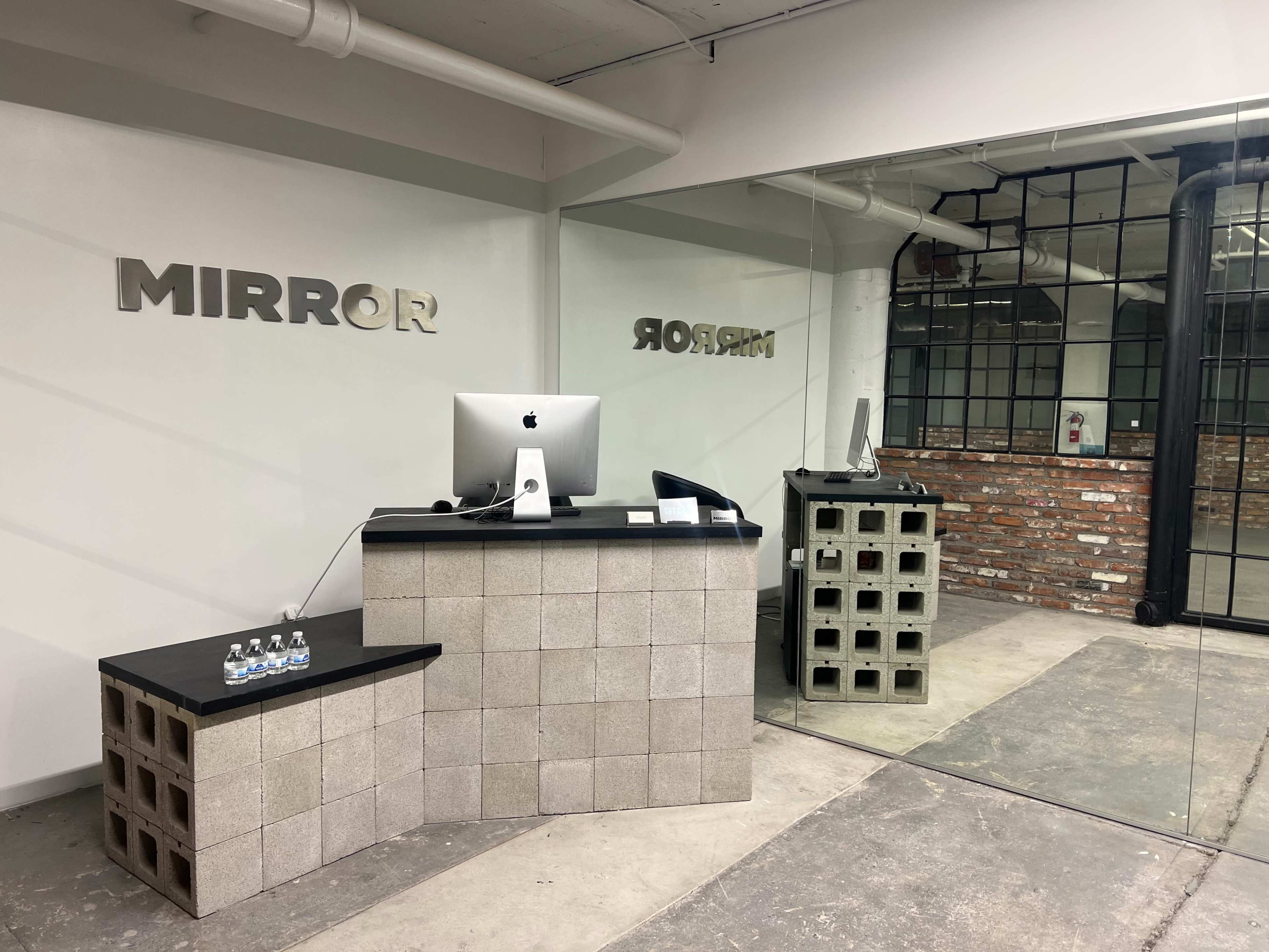The image shows a modern reception area with a desk made of concrete blocks, an iMac on the counter, and a large mirror along one wall.