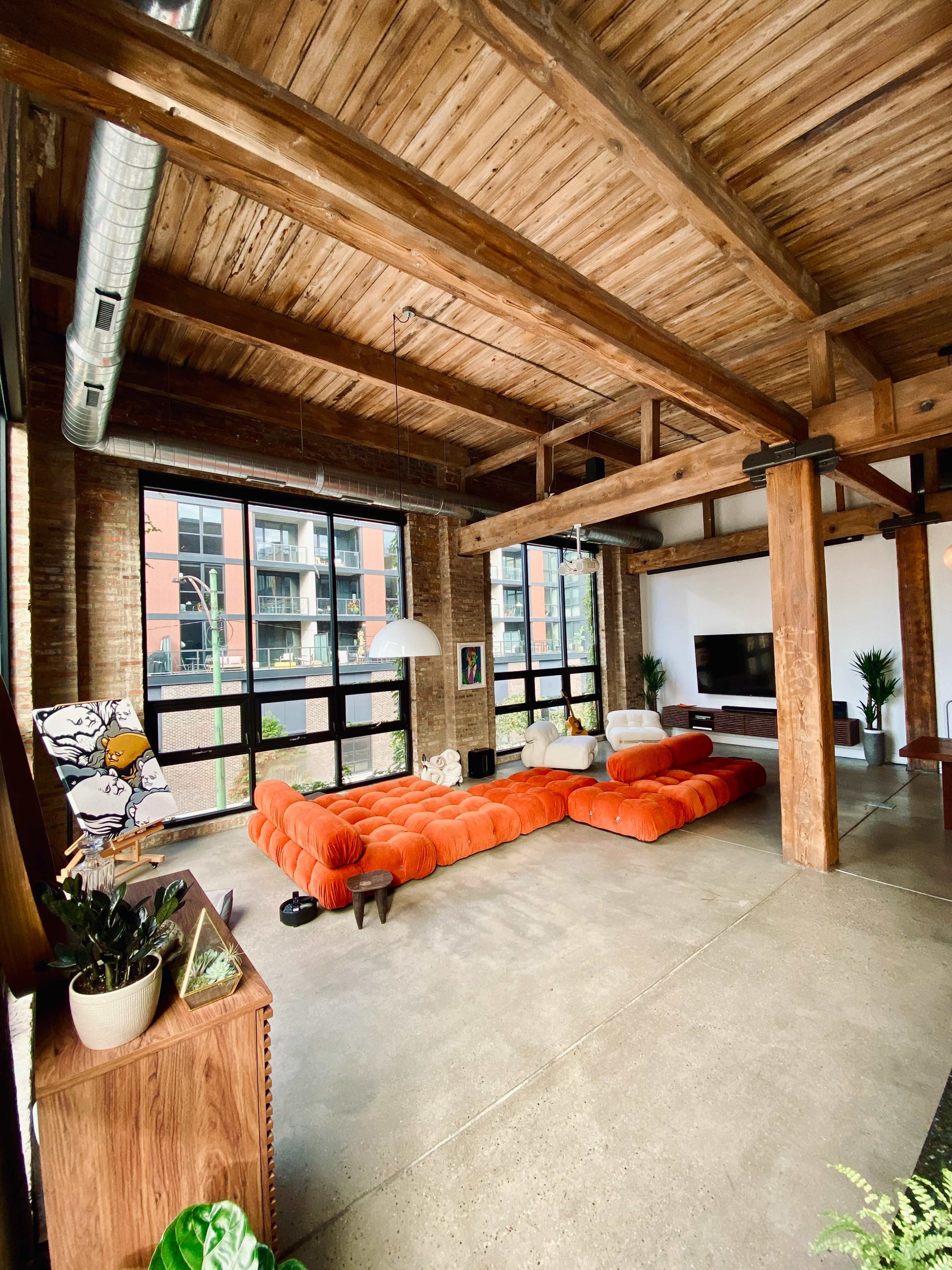 The spacious living area features a large orange sectional sofa, exposed wooden beams, and large windows that provide natural light.