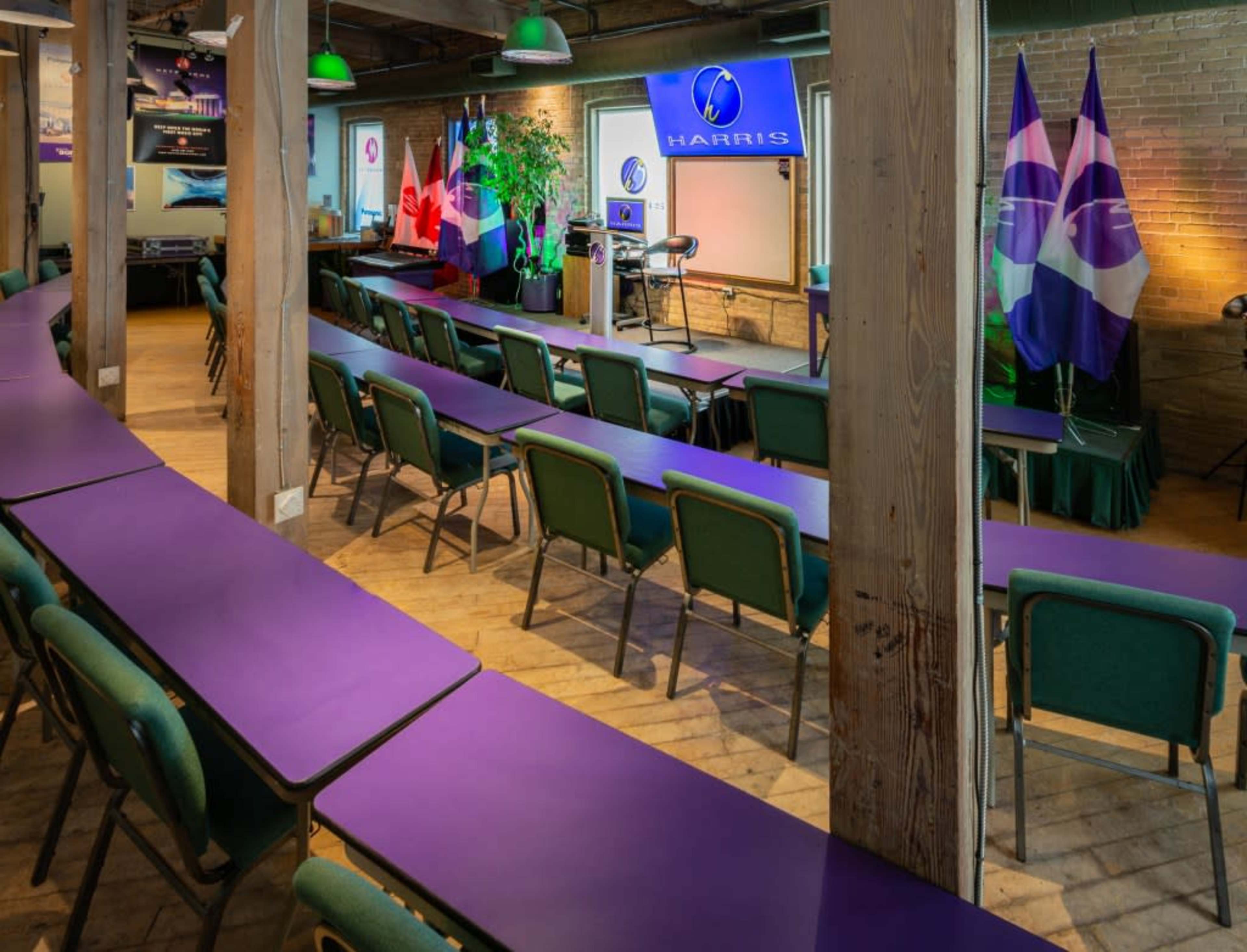A conference room set up with purple tables, green chairs, and flags, including the Canadian flag, arranged in front of a podium and screen displaying the word "HARRIS."