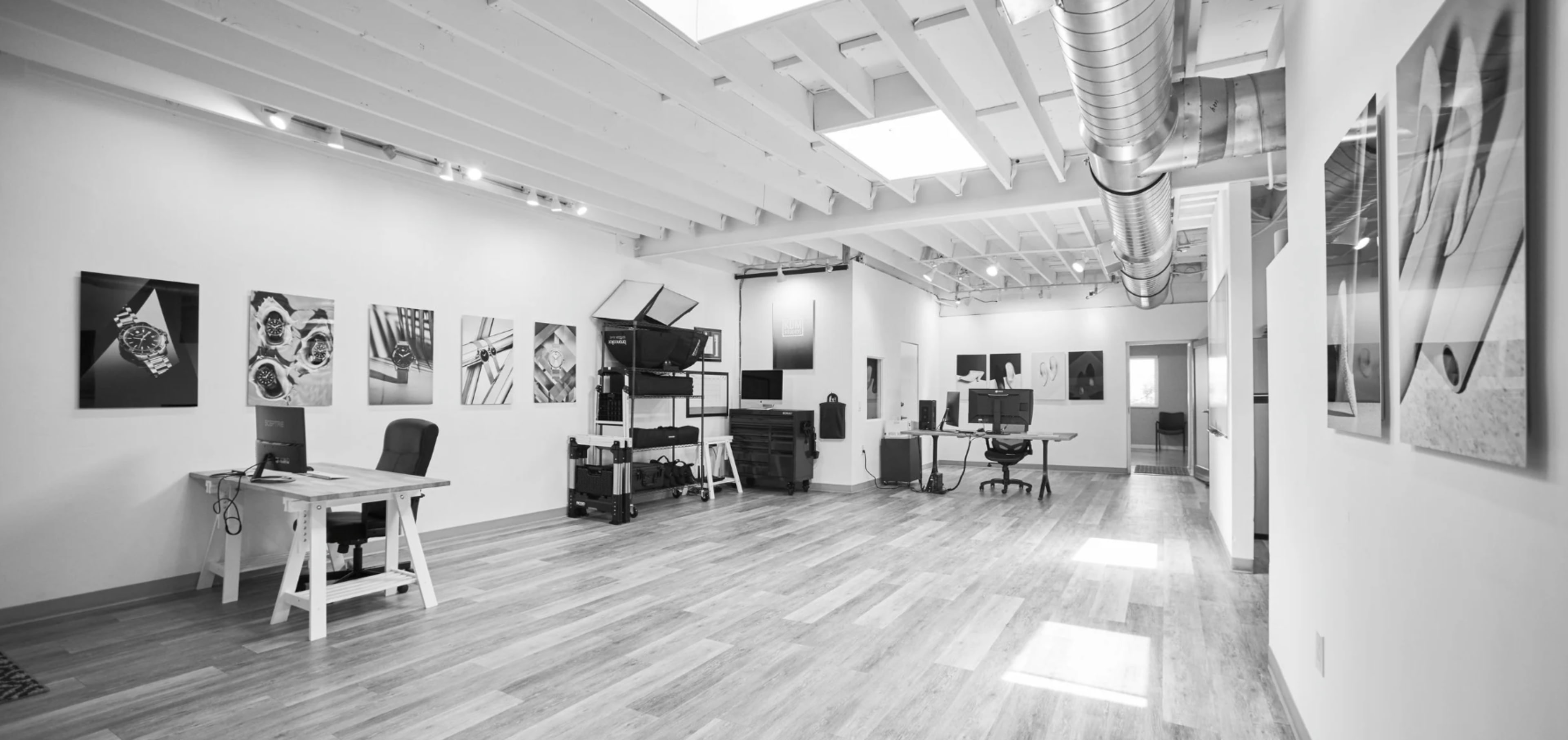 The image shows a spacious, minimalist office with wooden floors, white walls, and several pieces of artwork displayed on the walls.