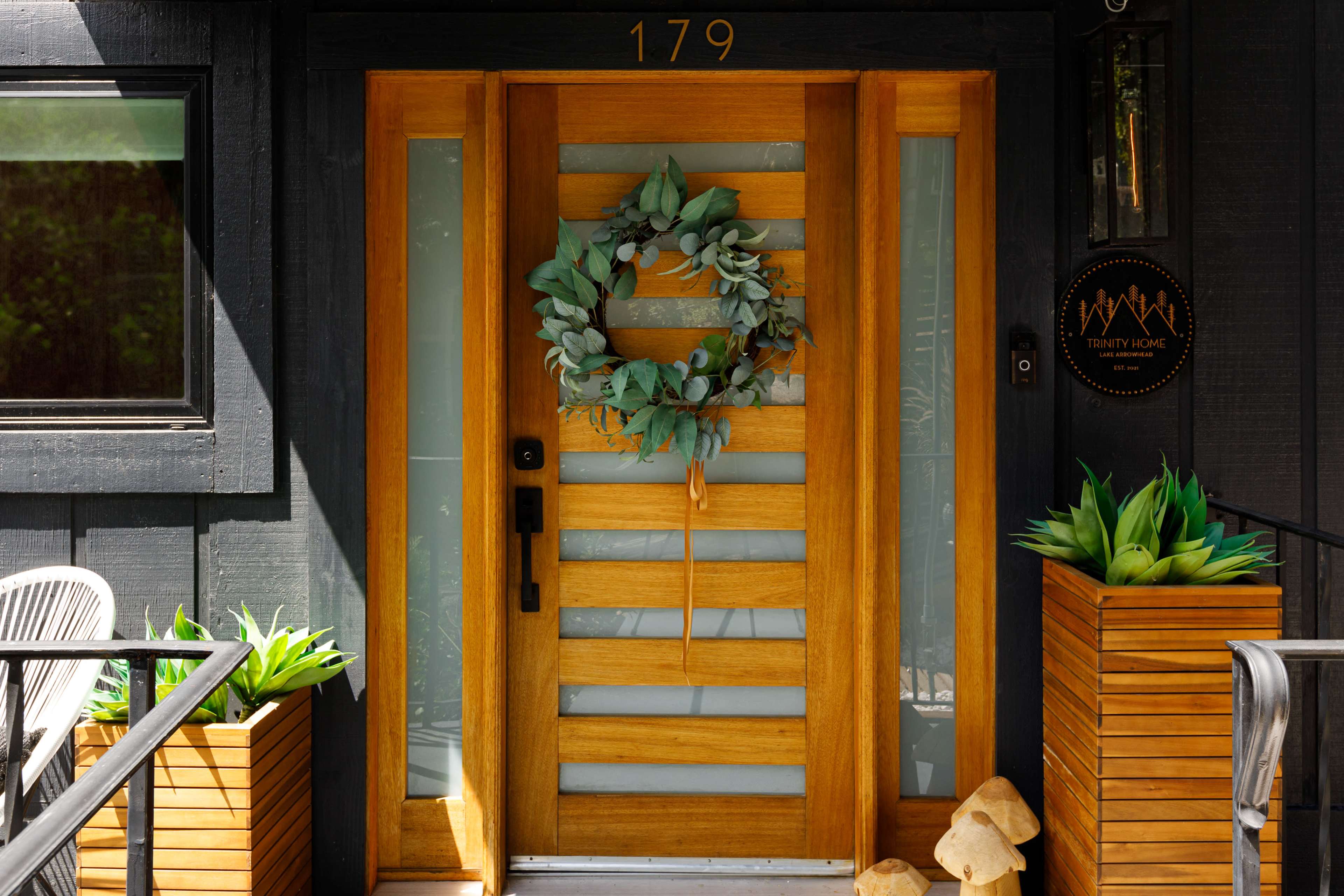 A modern wooden door with a horizontal slat design, adorned with a eucalyptus wreath, and flanked by decorative planters.