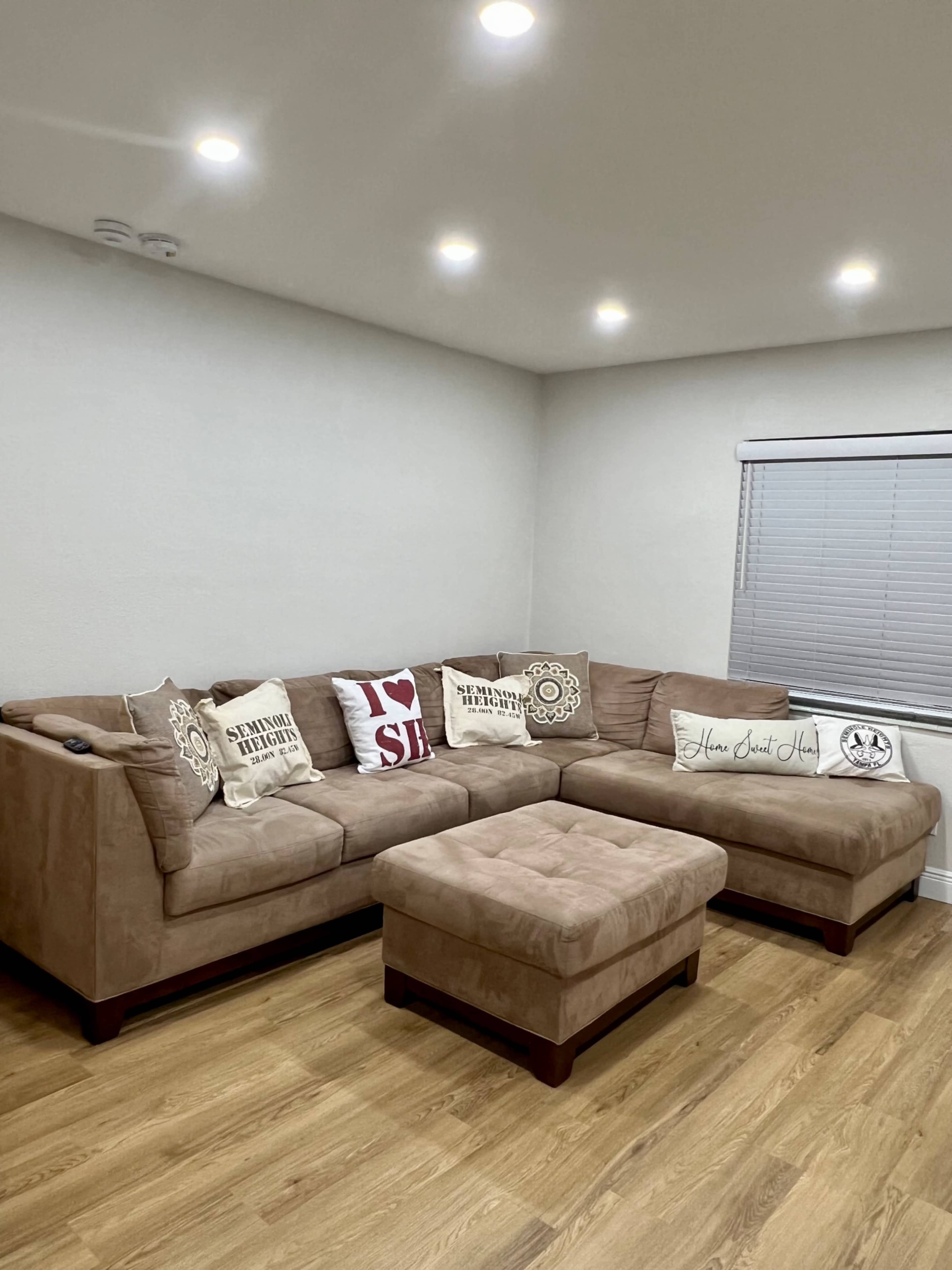 A light-colored sectional sofa with decorative pillows and an ottoman in a brightly lit room with wooden flooring.