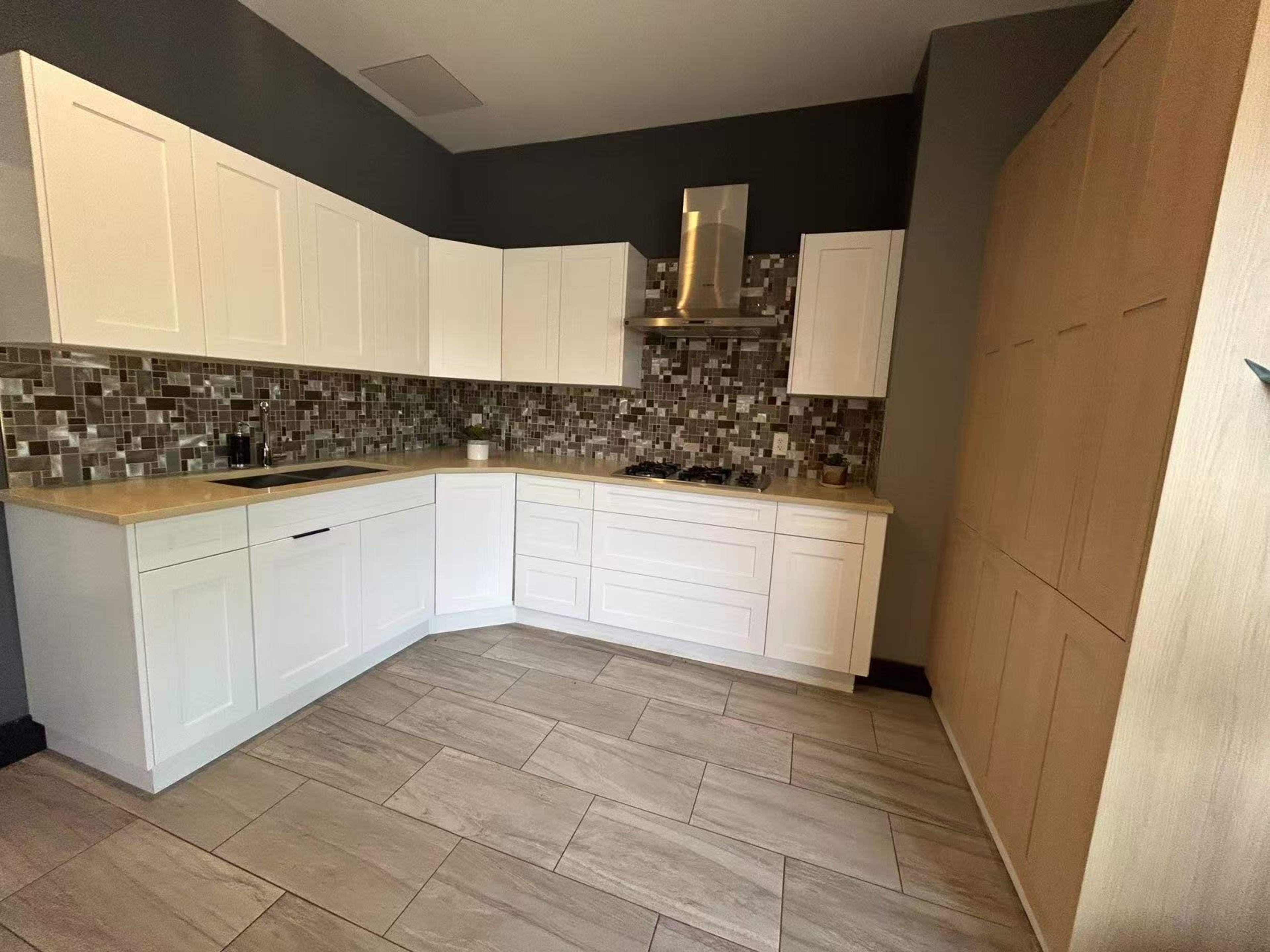 The image shows a modern kitchen with white cabinetry, a tiled backsplash in shades of gray and brown, and light-colored tile flooring.
