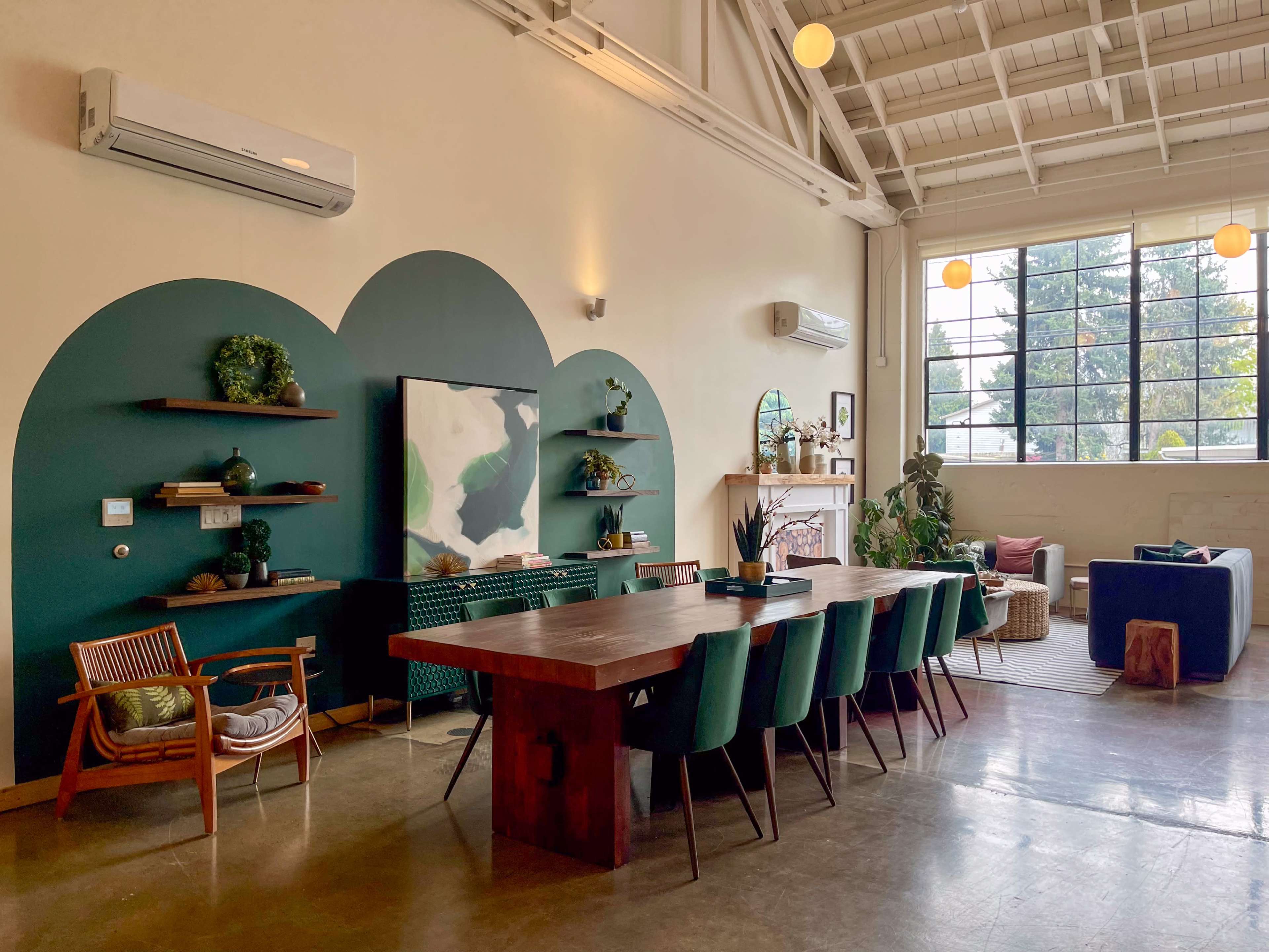 The image shows a spacious interior featuring a long wooden dining table with green chairs, a large wall-mounted TV, and various decorative items, all under a high ceiling with large windows allowing natural light.