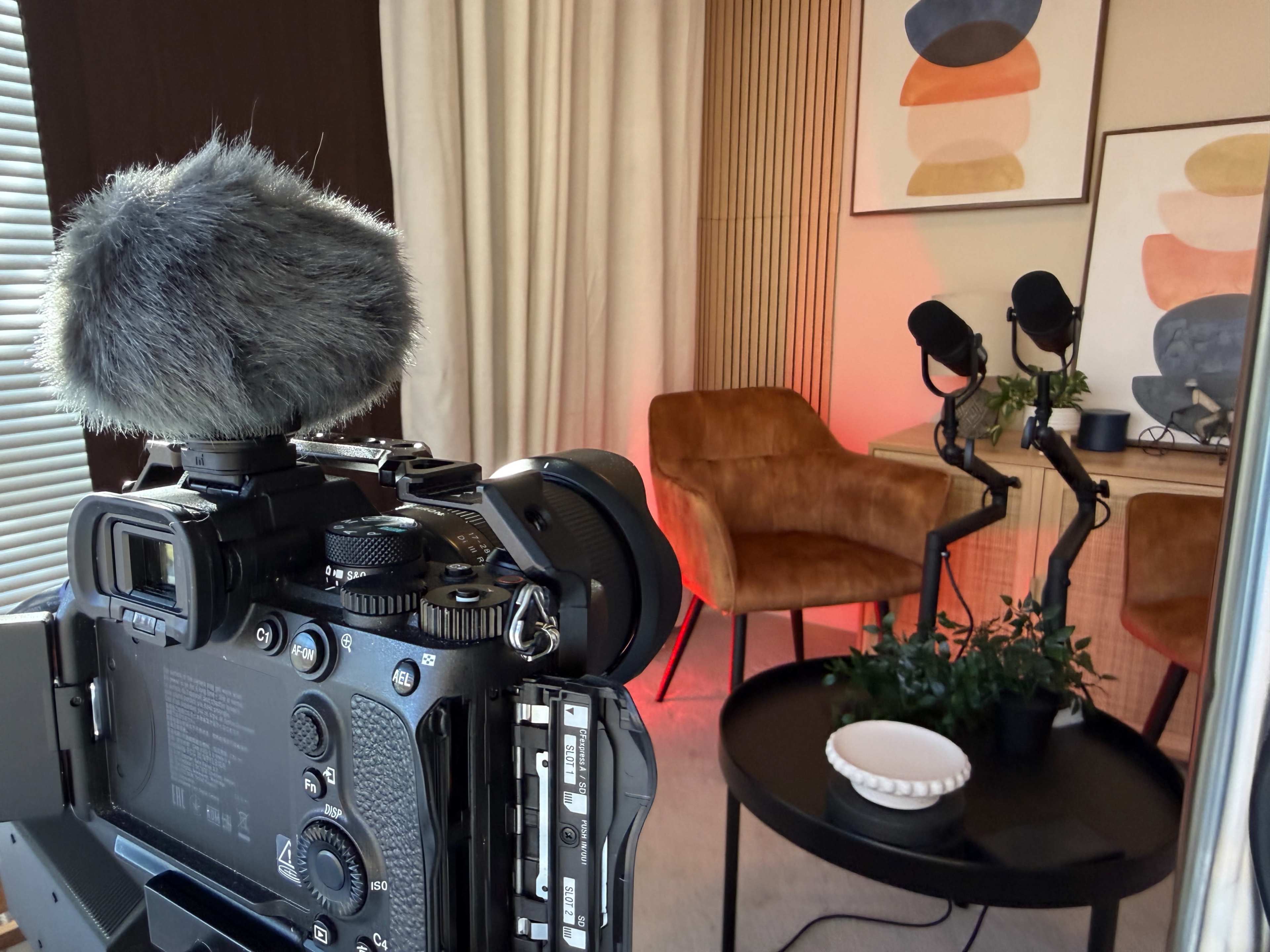 A camera with a microphone is positioned in front of two orange chairs and a table with a plant and bowl in a well-lit room decorated with abstract art.
