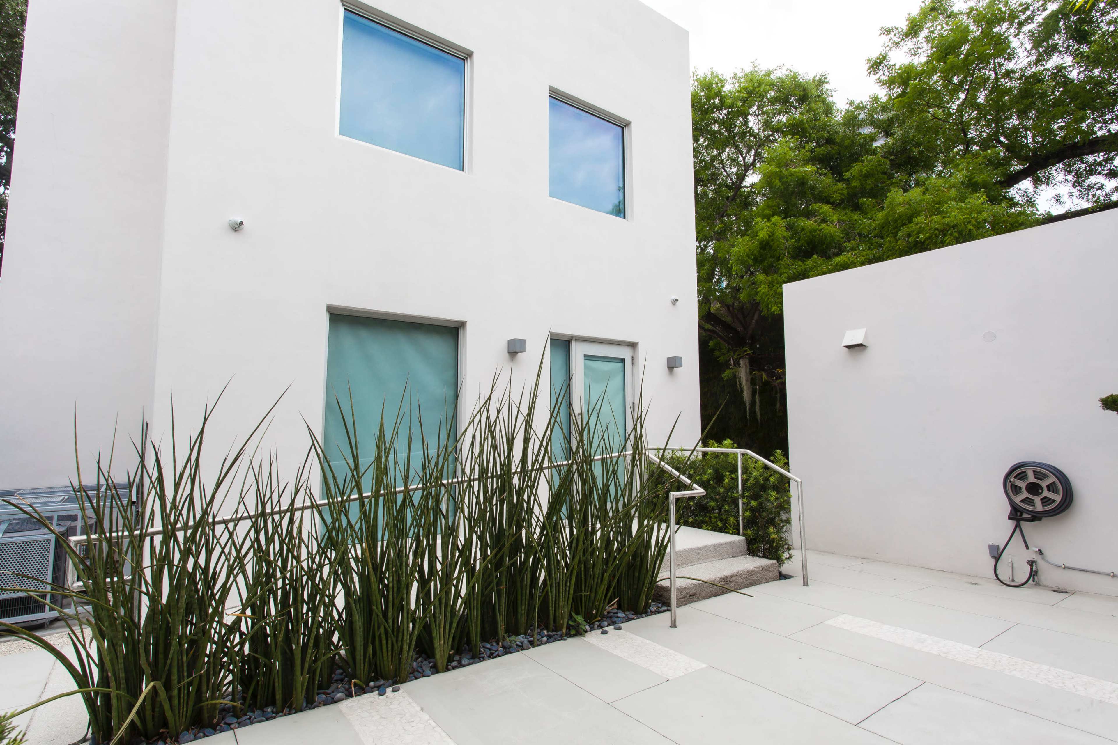 The image shows a modern, two-story white building surrounded by tall green plants and a gray stone pathway.