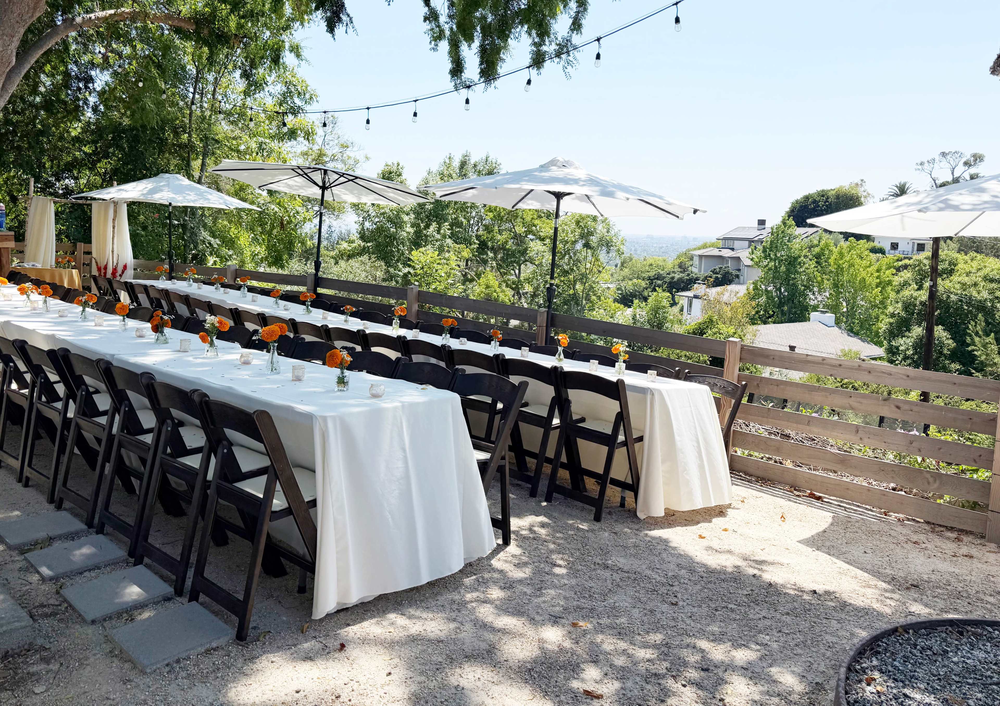 A long table with white linens and orange flower centerpieces is set outdoors under white umbrellas, overlooking a landscape of trees and houses.