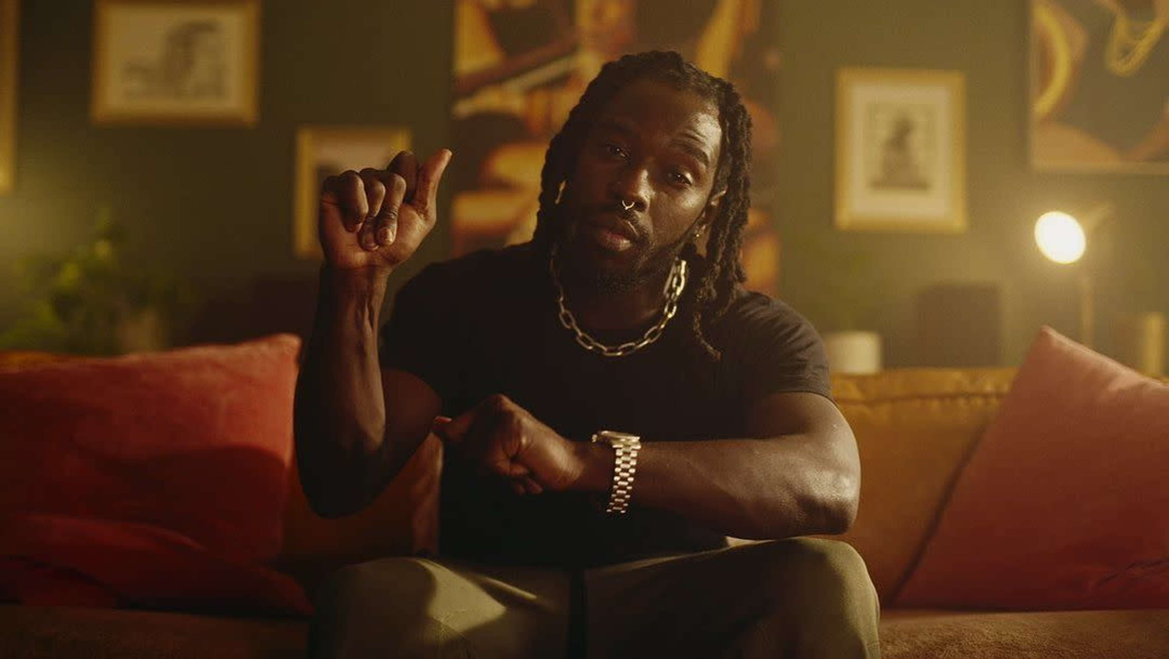 A man wearing a black shirt and silver jewelry gestures while seated on a couch in a warmly lit room decorated with framed artwork.