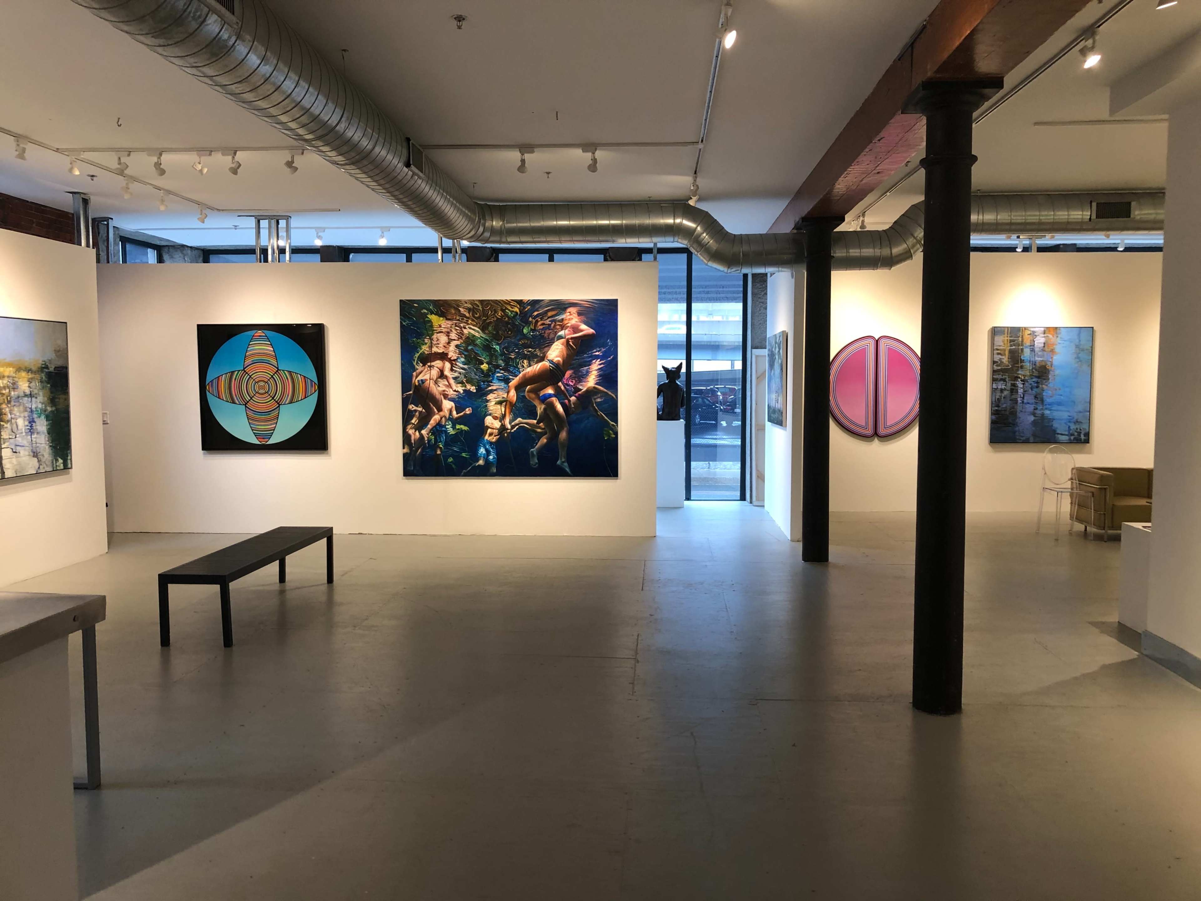 The image shows an art gallery with several colorful paintings displayed on white walls and a minimalist seating area in the background.