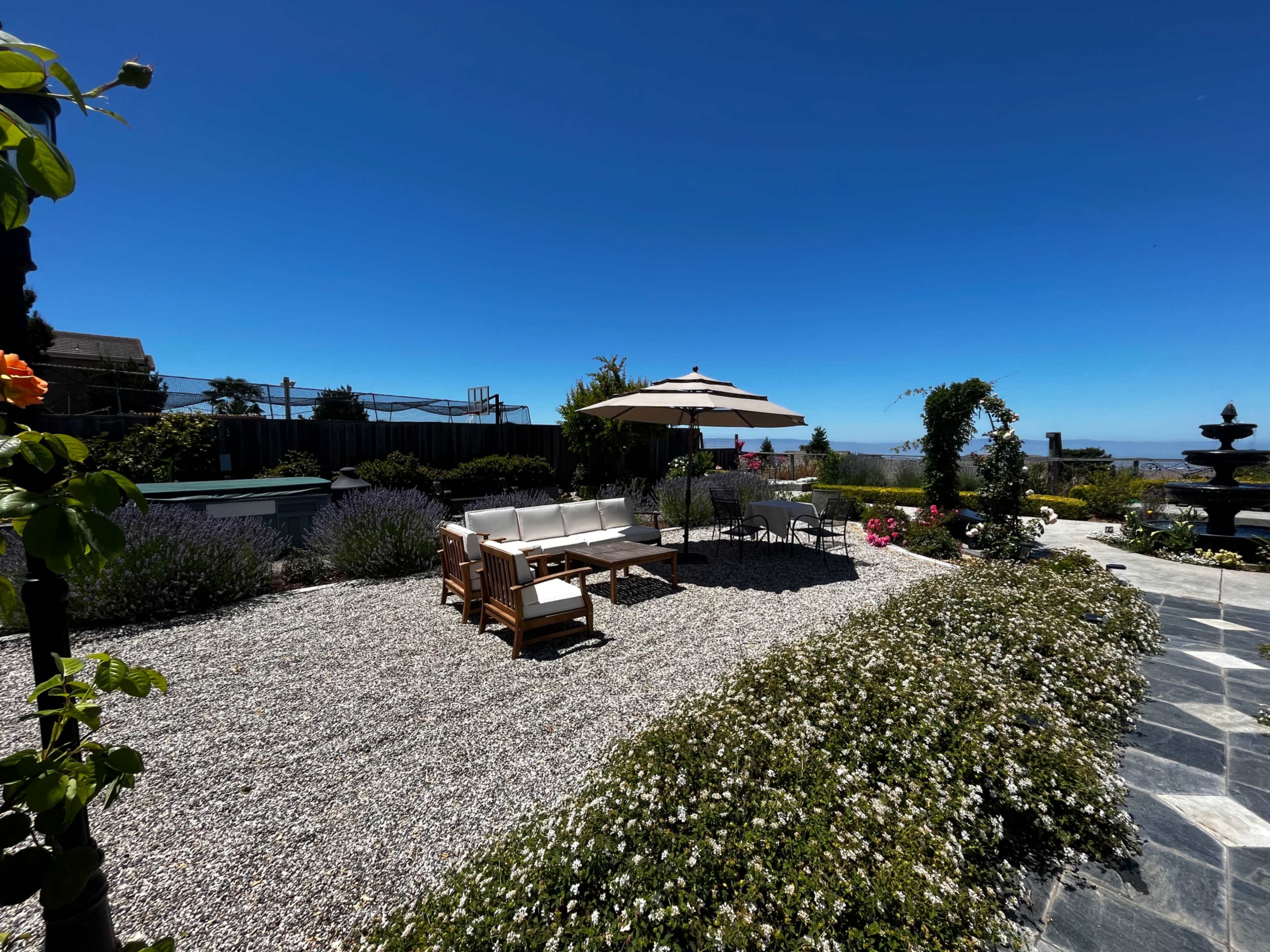A gravel patio features wooden seating and an umbrella, surrounded by blooming plants and a view of the sky.
