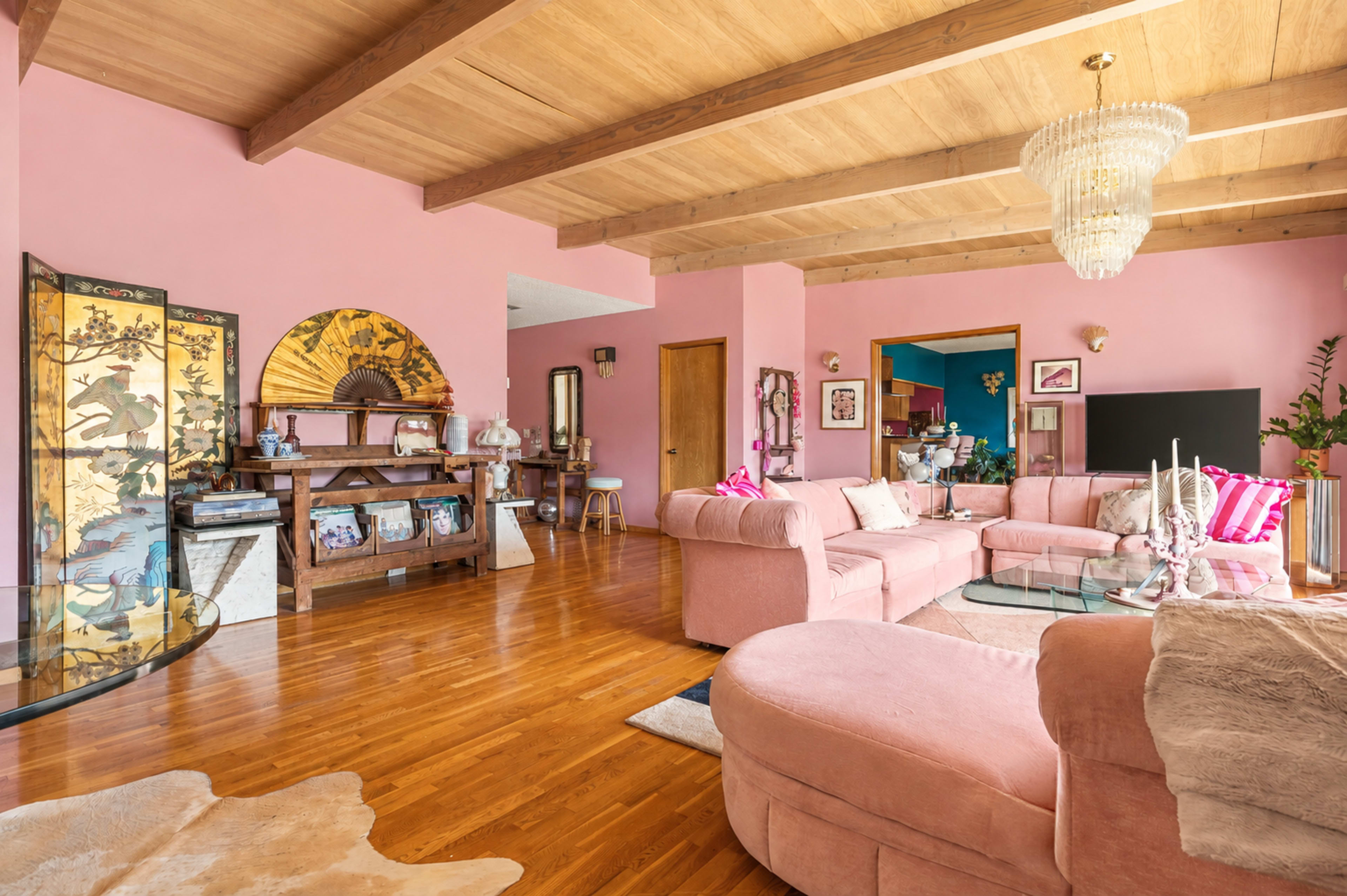 The image depicts a brightly colored living room featuring pink walls, wooden beams, a chandelier, and comfortable seating arranged around a glass coffee table.