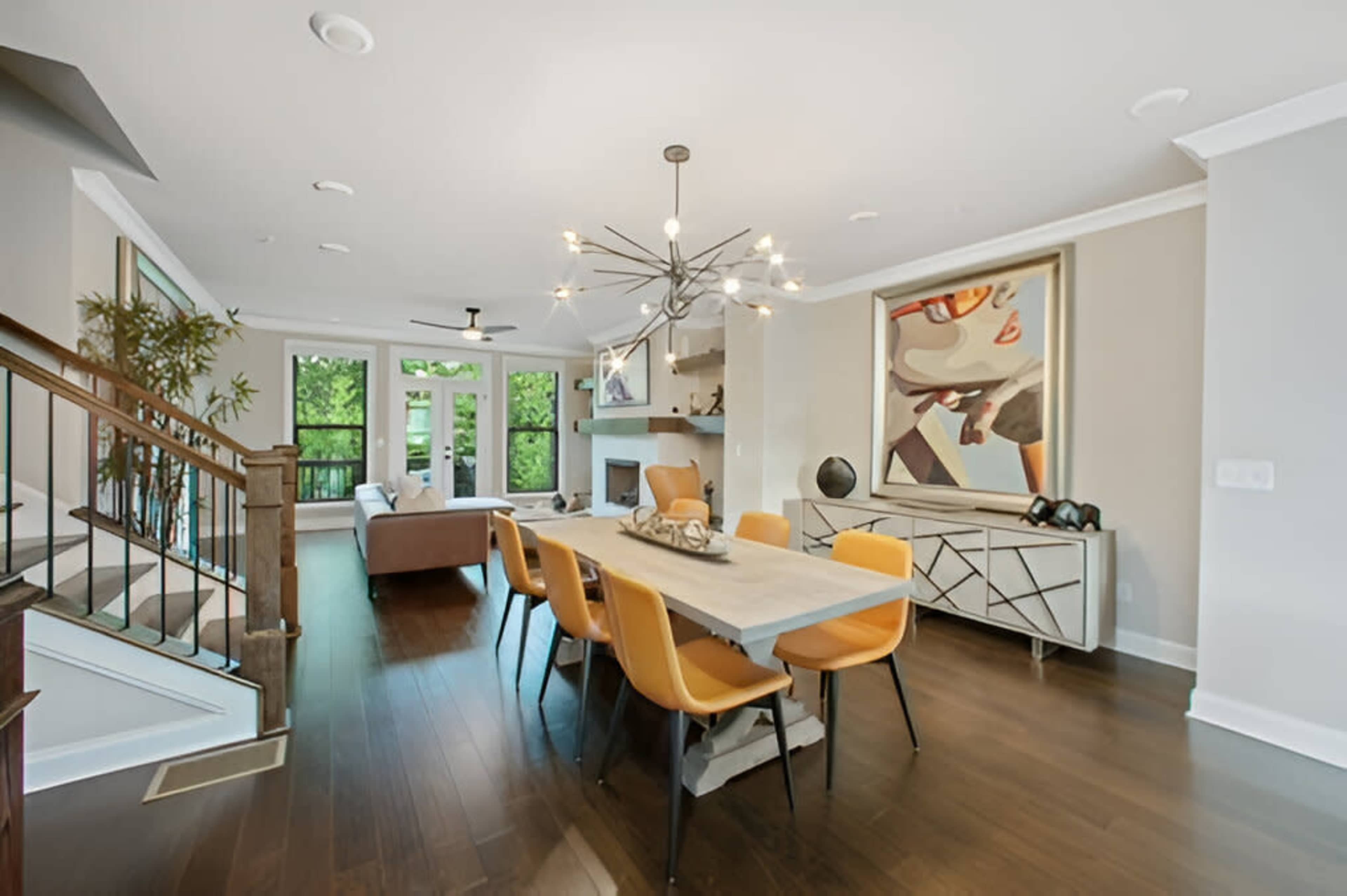 A modern dining area featuring a rectangular table with yellow chairs, a chandelier above, and an adjacent living space with a couch and large windows.