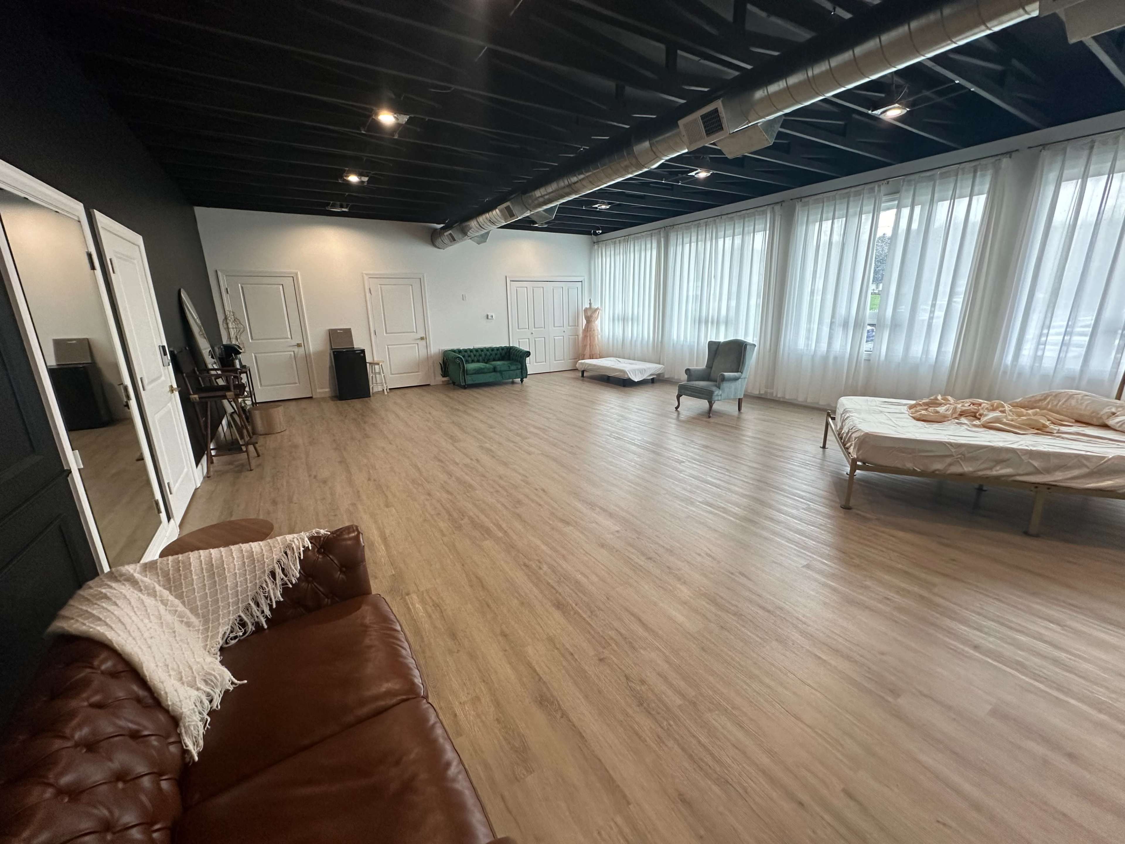 A spacious studio with wooden flooring, large windows covered by sheer curtains, and a mix of furniture including a green couch, a bed, and a chair.
