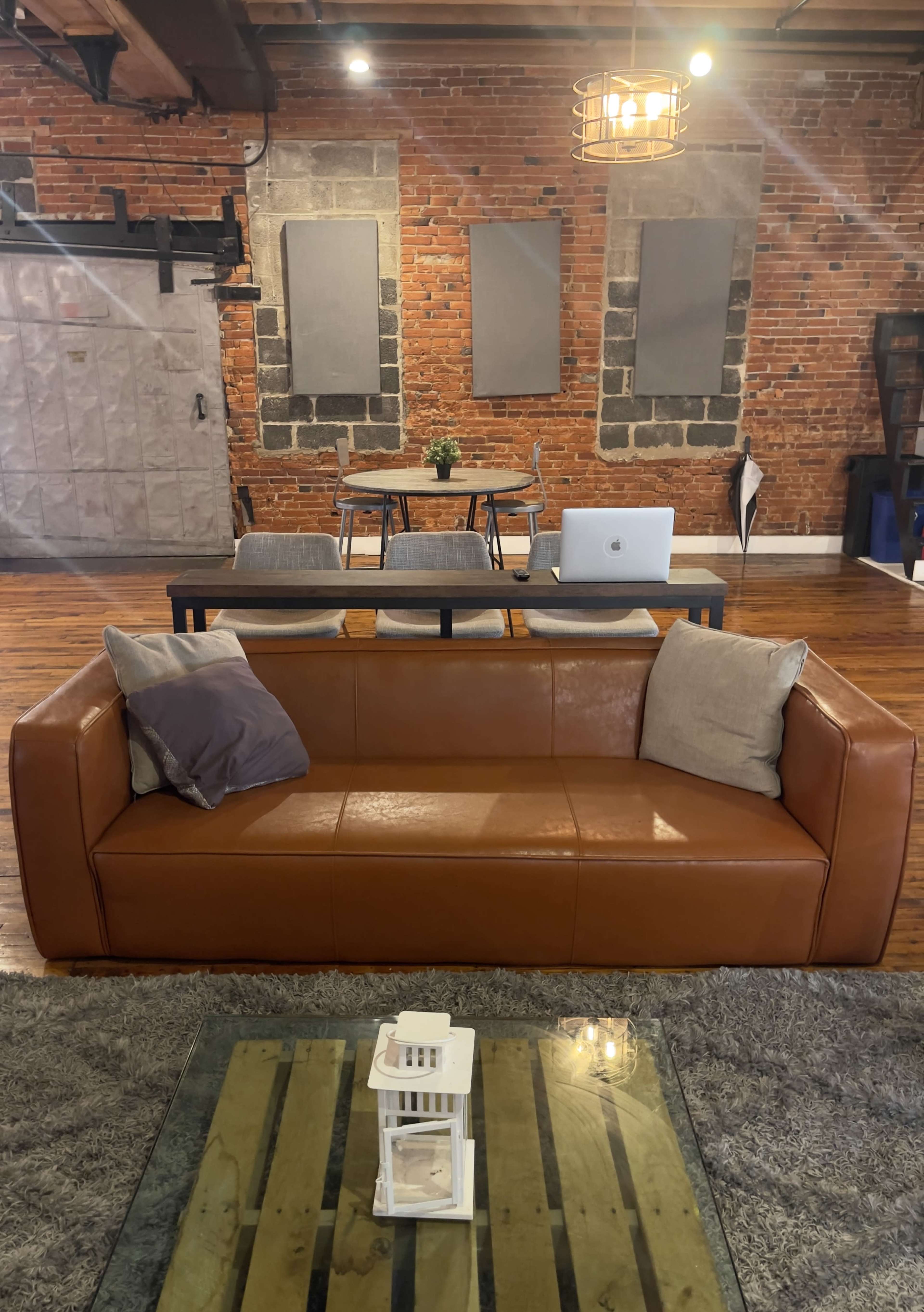 A brown leather sofa sits in the foreground of a room featuring exposed brick walls, a modern dining table with chairs in the background, and a laptop on the table.