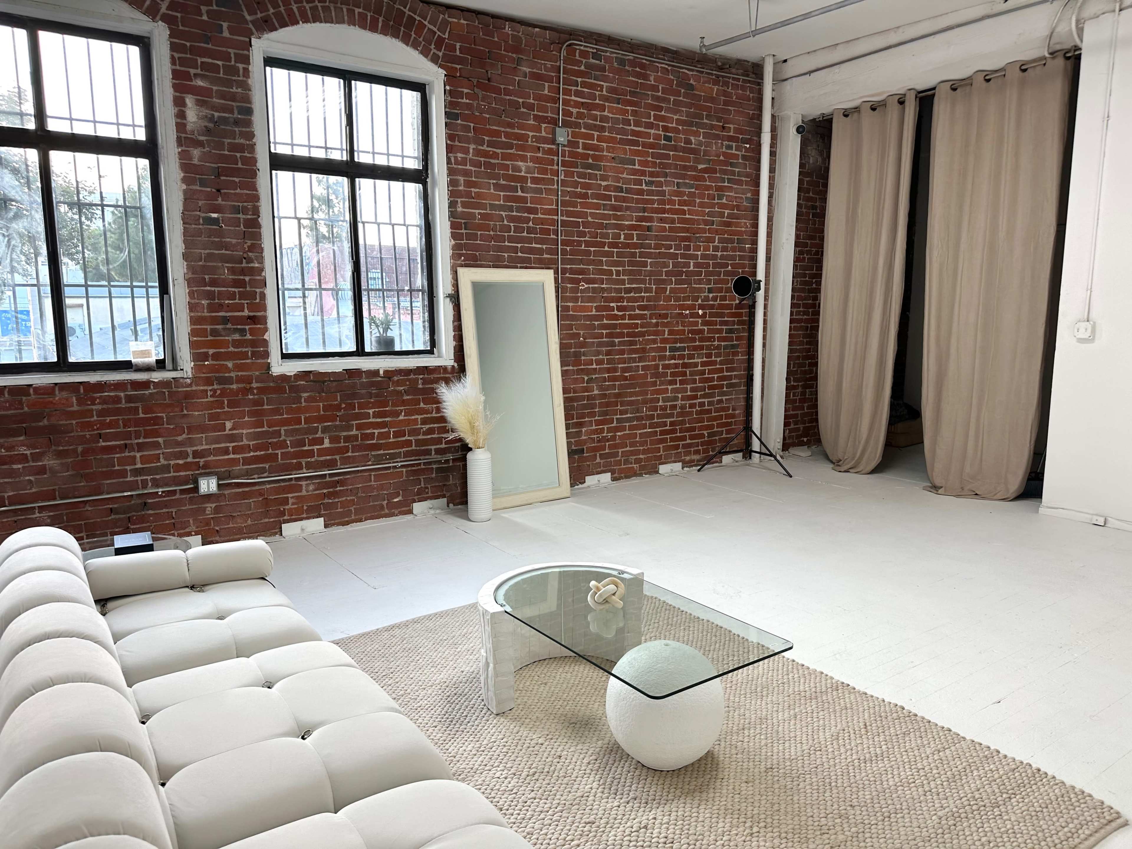 A minimalist interior space featuring a white tufted sofa, a round glass coffee table, and exposed brick walls with large windows.
