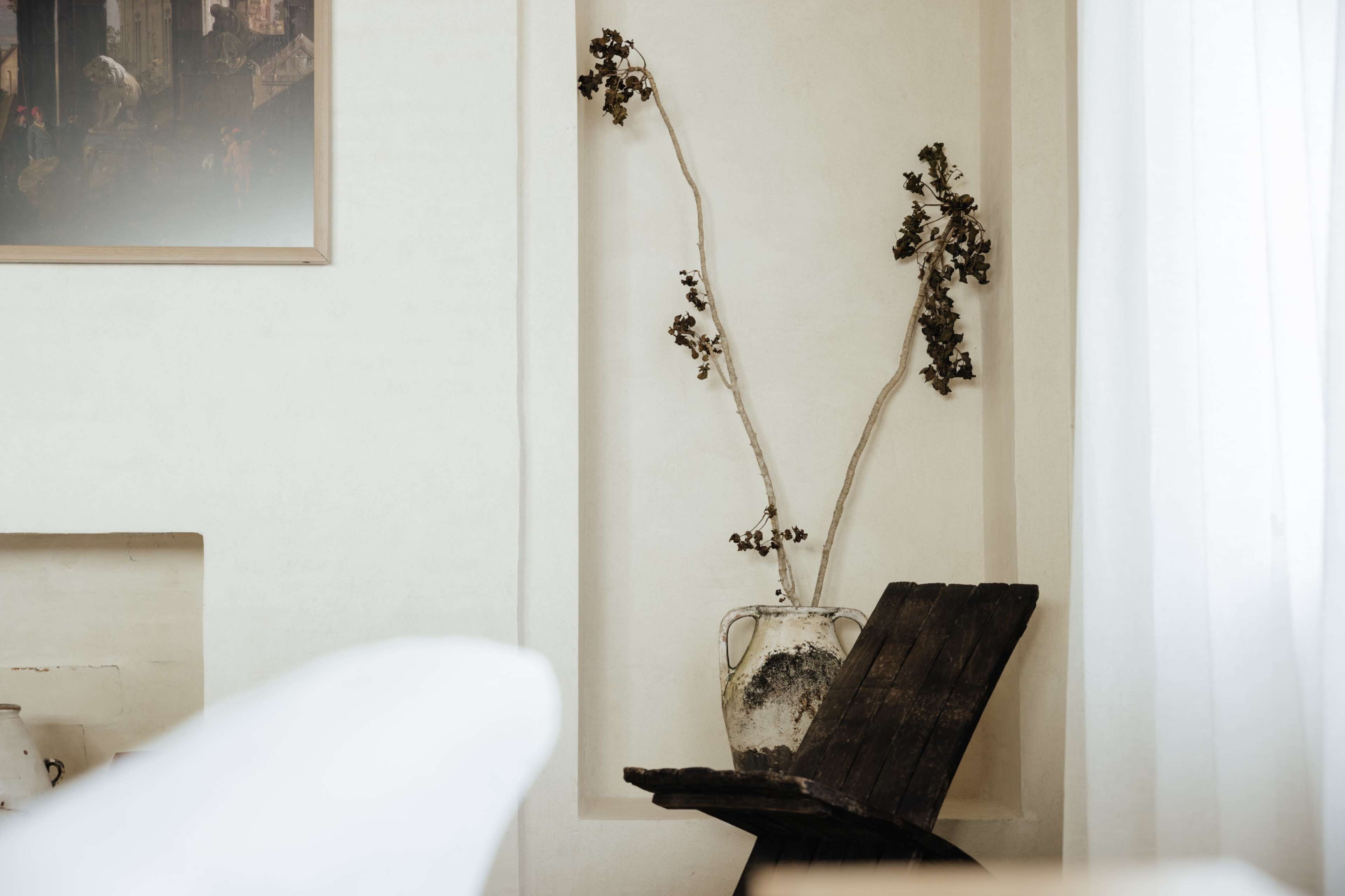 A rustic wooden chair with a simple design holds a weathered vase containing dry branches against a textured wall.