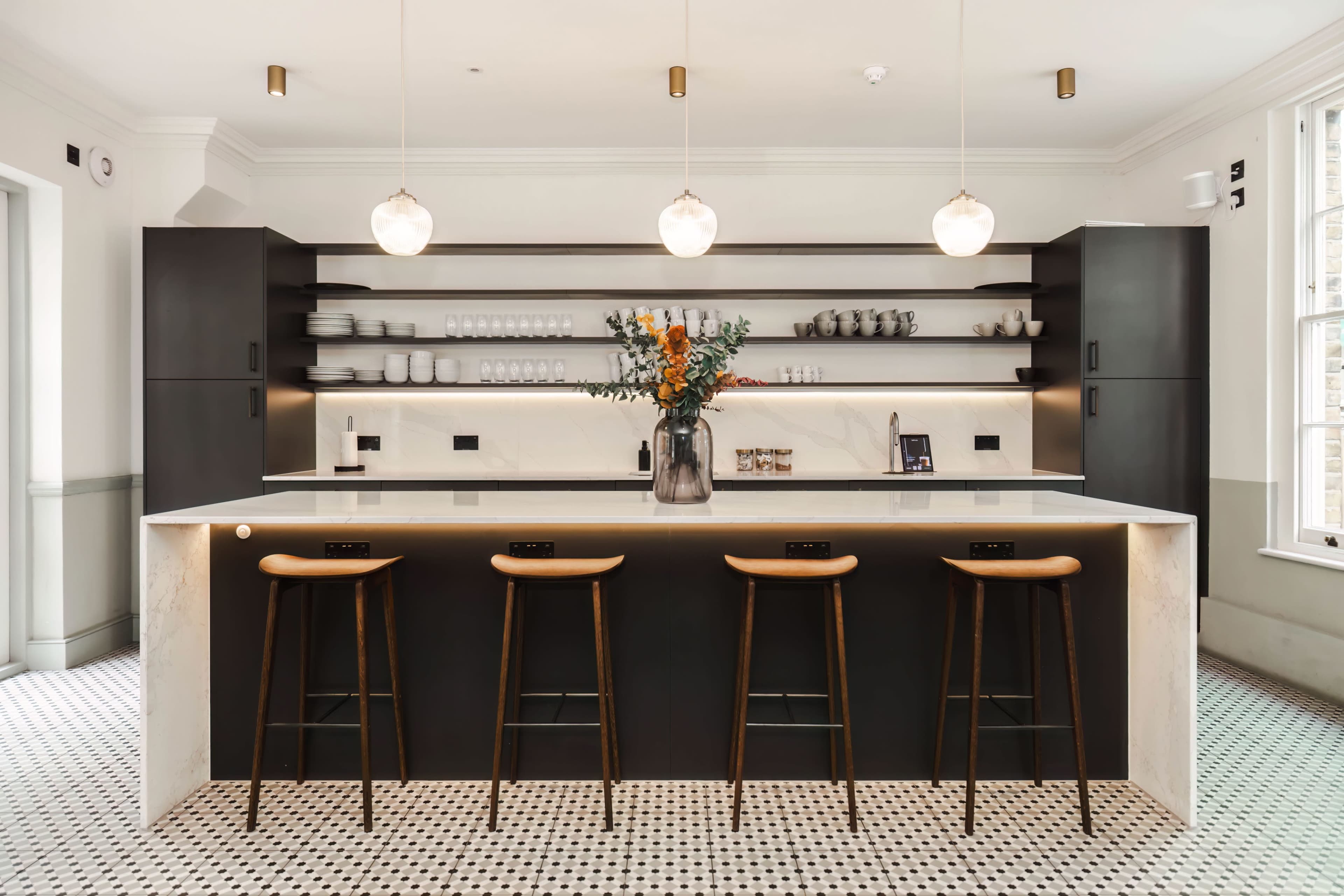 A modern kitchen features a central island with three wooden bar stools and a floral arrangement, surrounded by dark cabinetry and open shelves.
