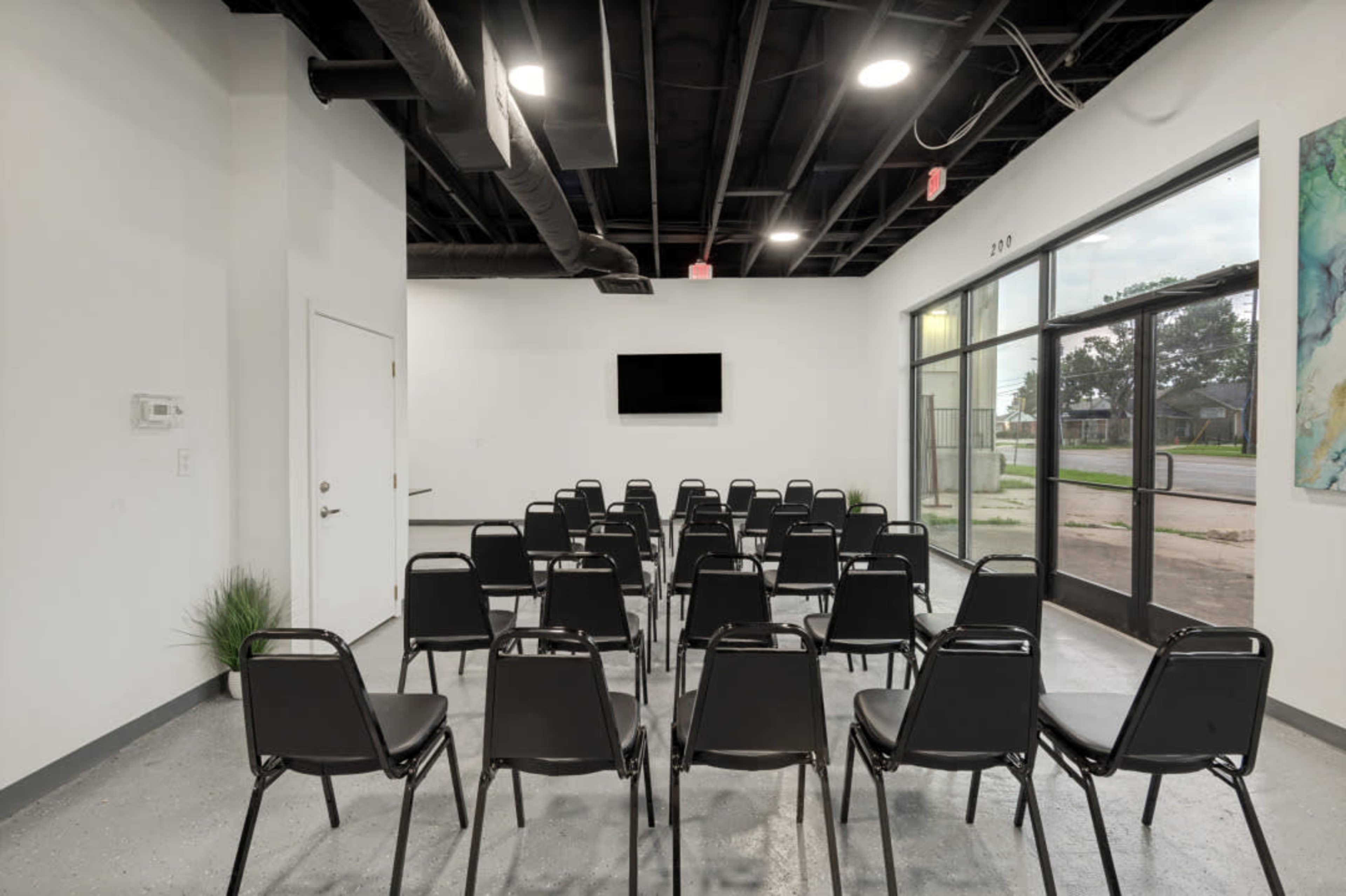 The image shows a minimalist meeting room with rows of black chairs facing a wall-mounted screen.