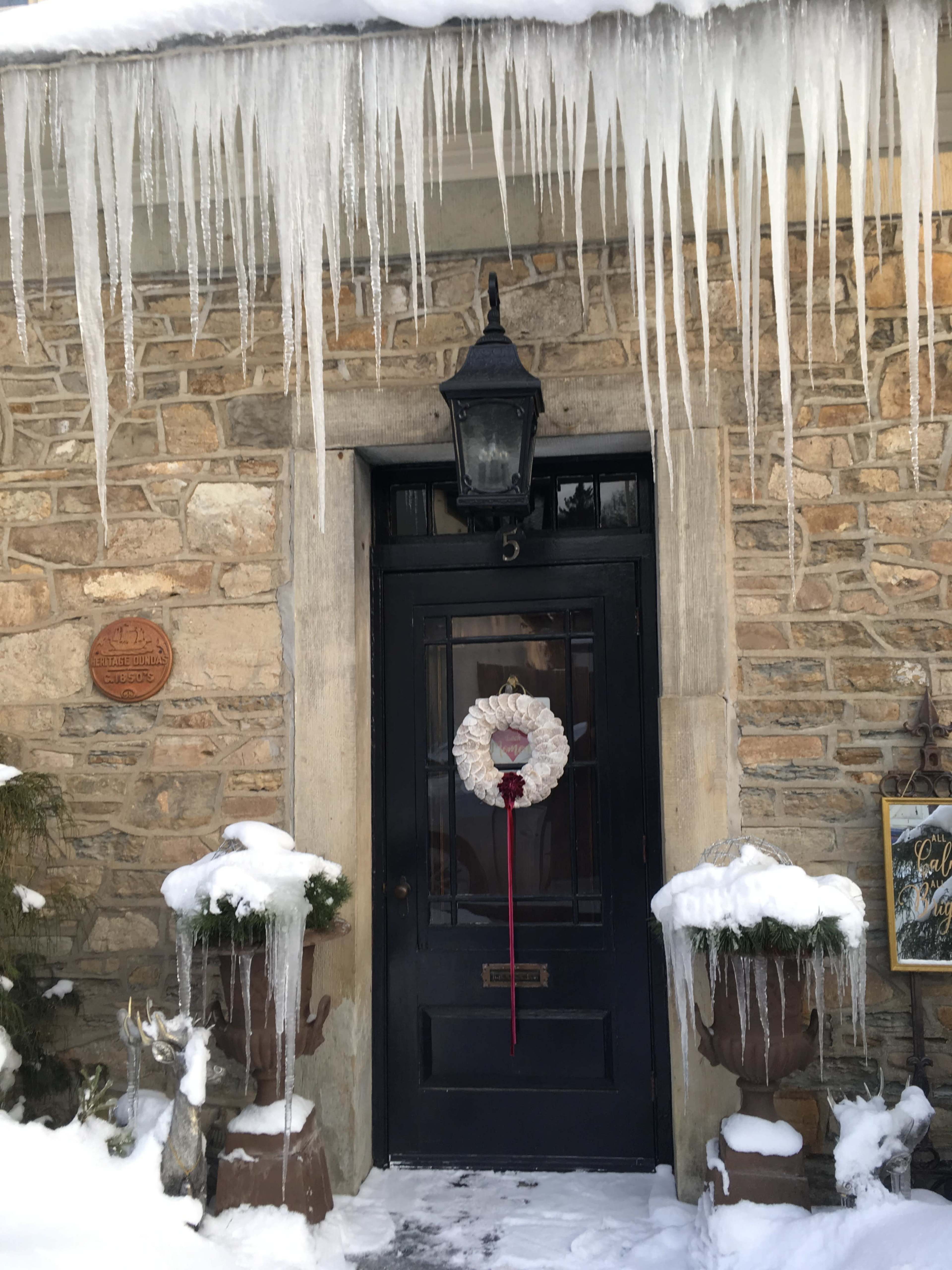 A black front door adorned with a heart-shaped wreath is framed by snow-covered planters and icicles hanging from a stone wall.