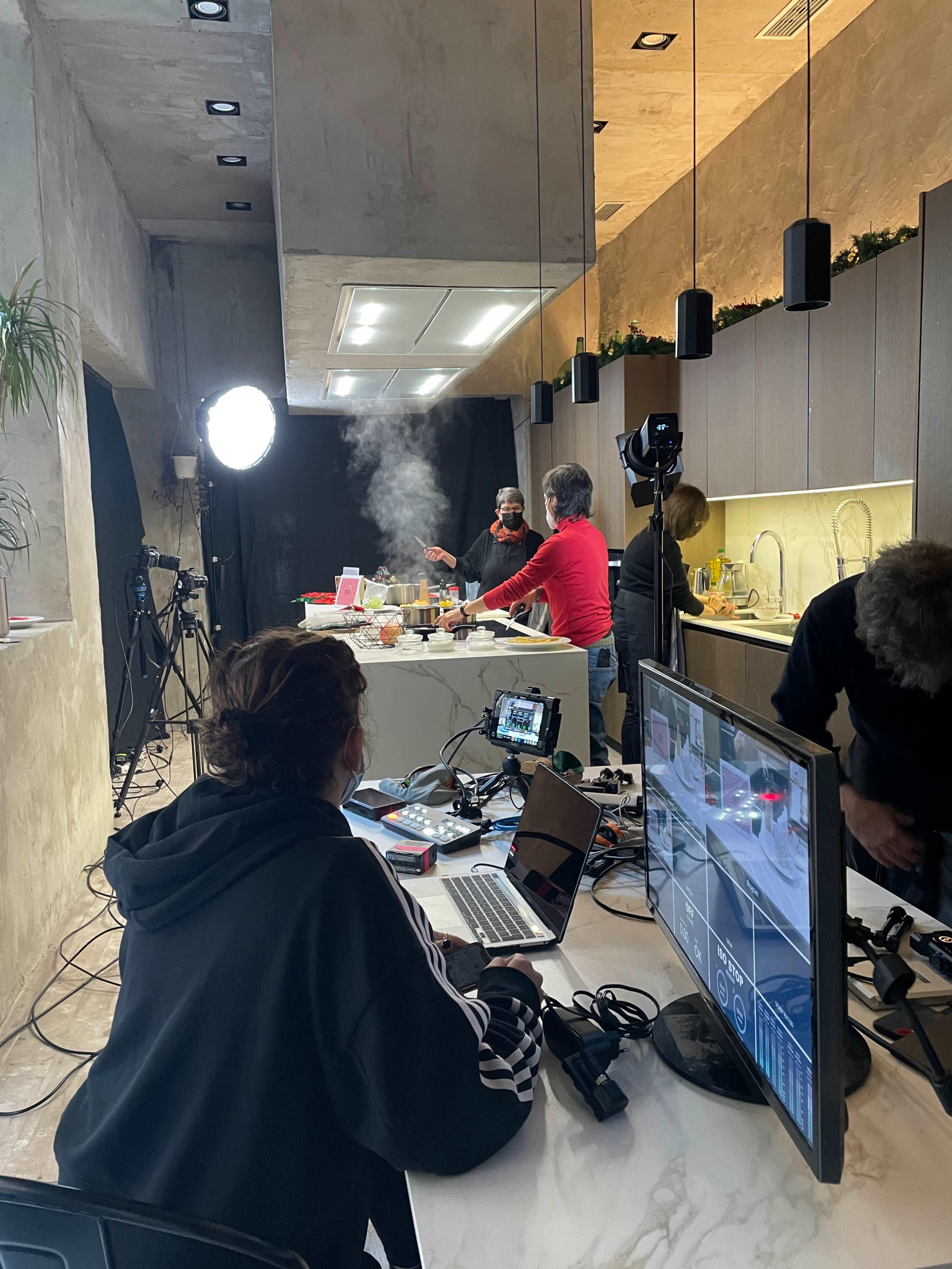 A group of people prepares food in a modern kitchen while a person operates a camera setup in the foreground.