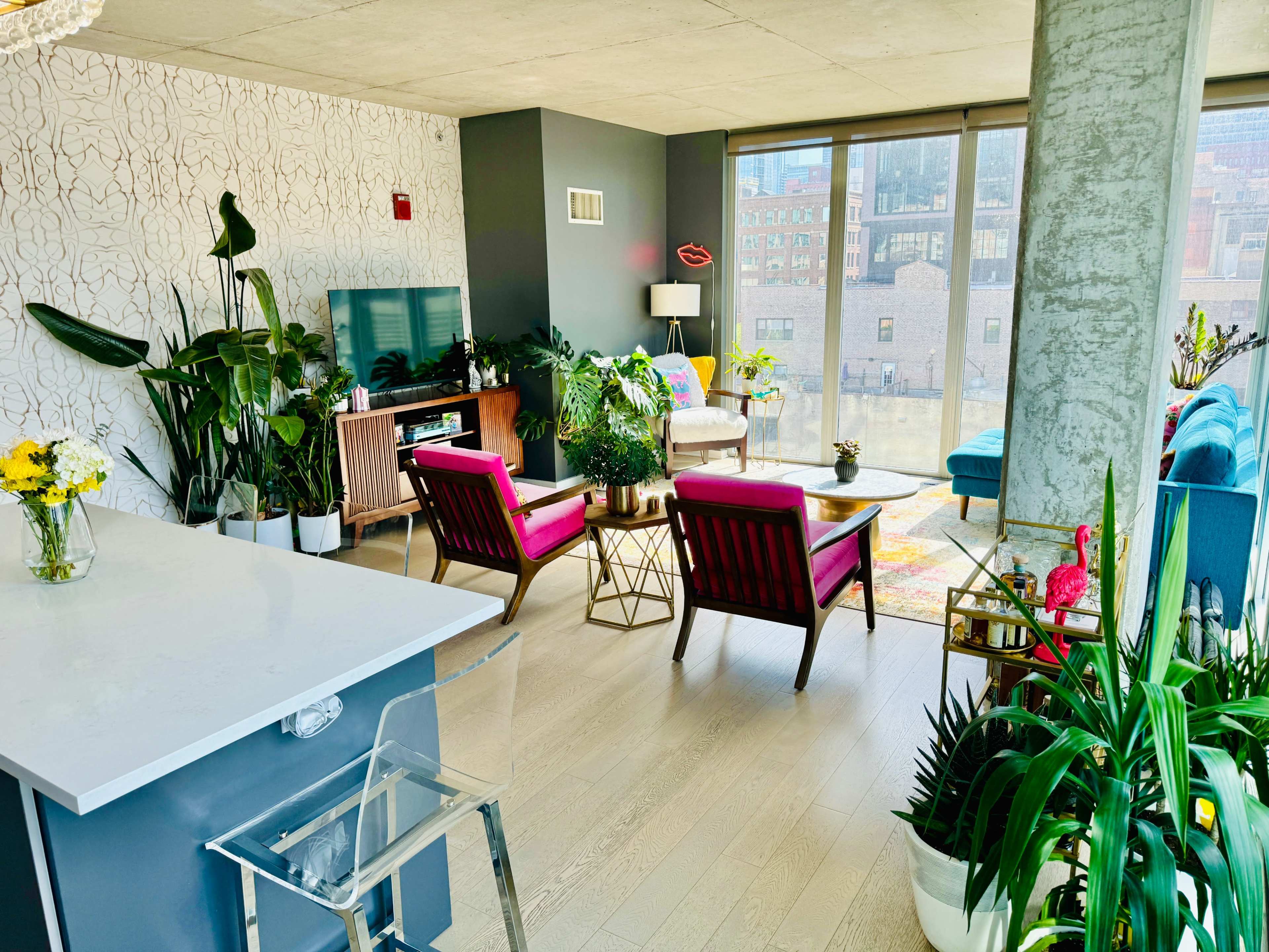 The scene features a modern living room filled with various plants, colorful furniture, and large windows allowing natural light to fill the space.