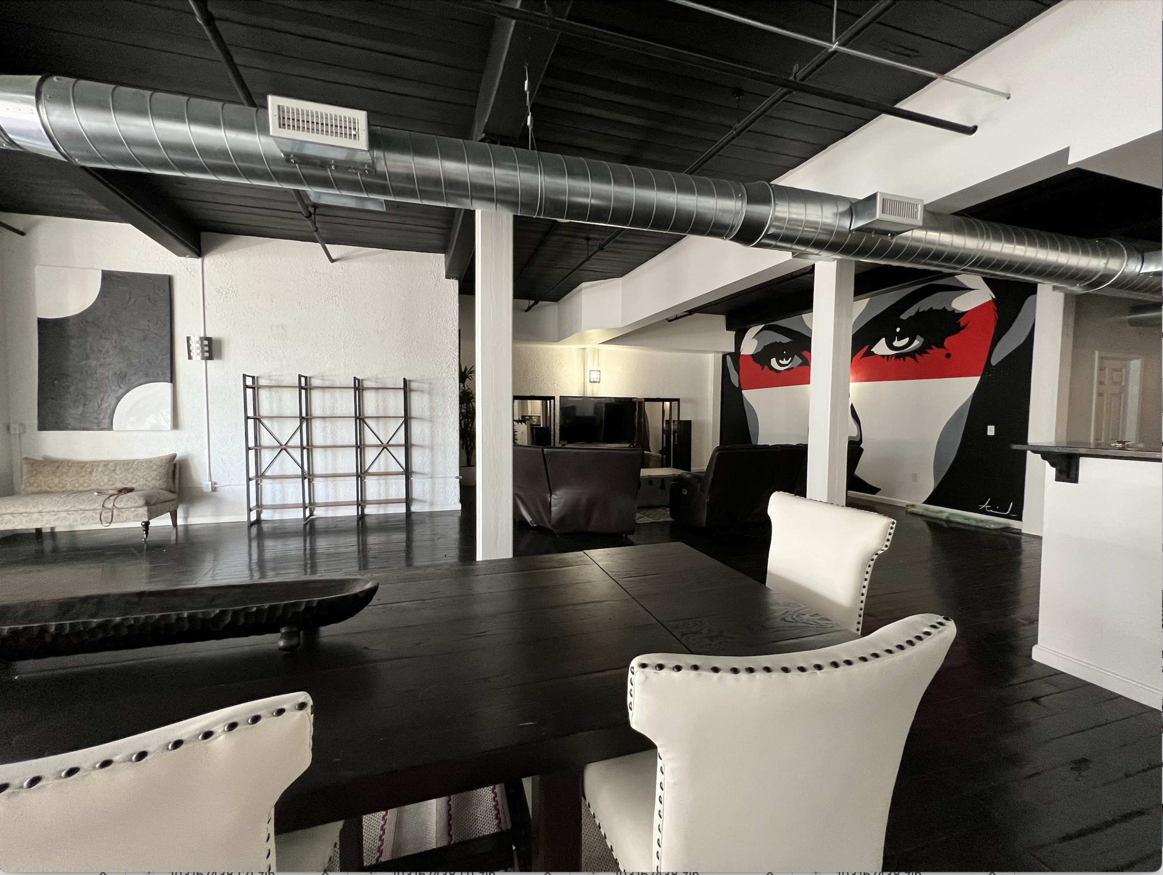 A spacious interior with a dining table and chairs in the foreground, a large wall mural of a face in the background, and various furniture pieces in an open living area.