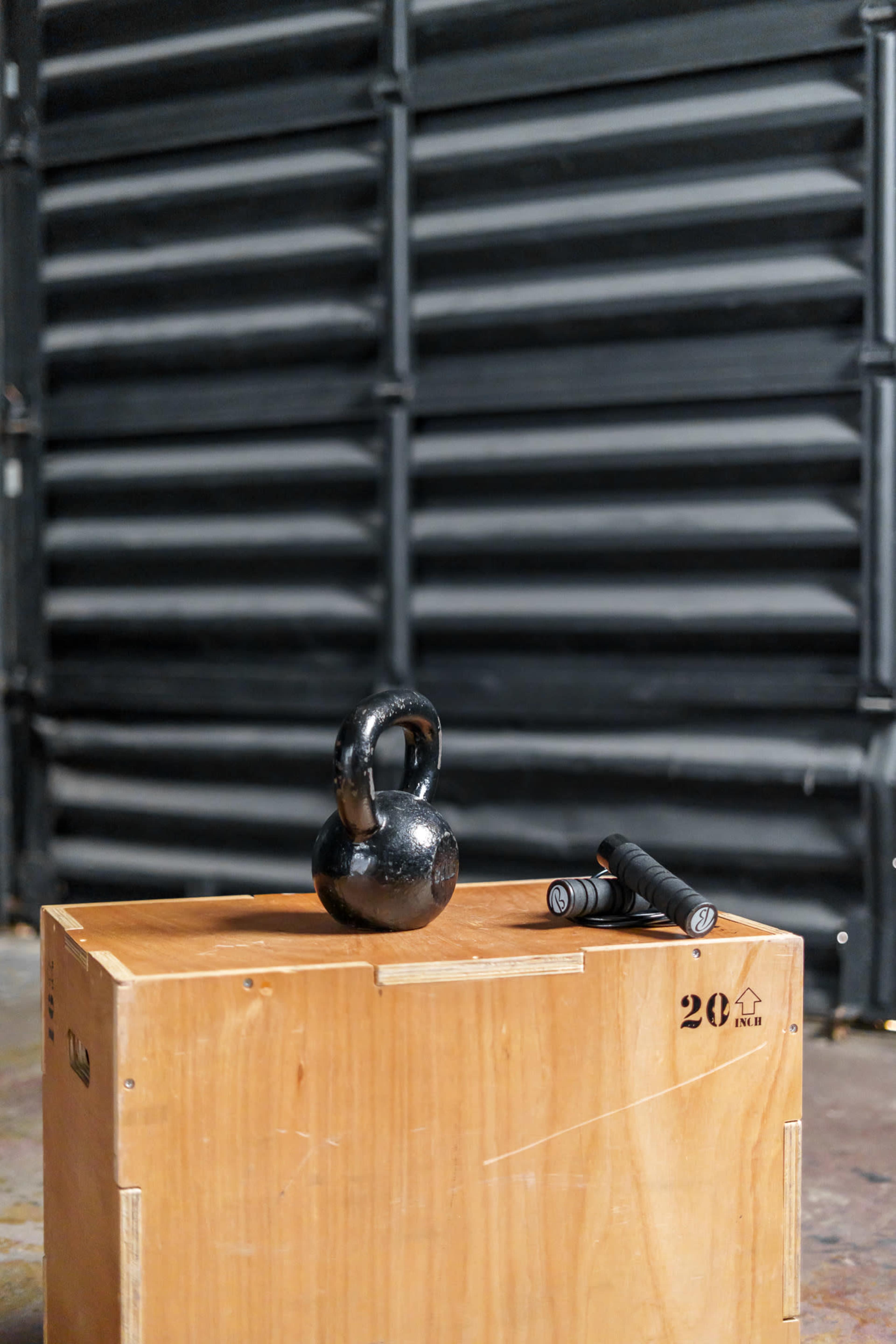 A black kettlebell and a pair of dumbbells rest on a wooden box against a dark, corrugated background.