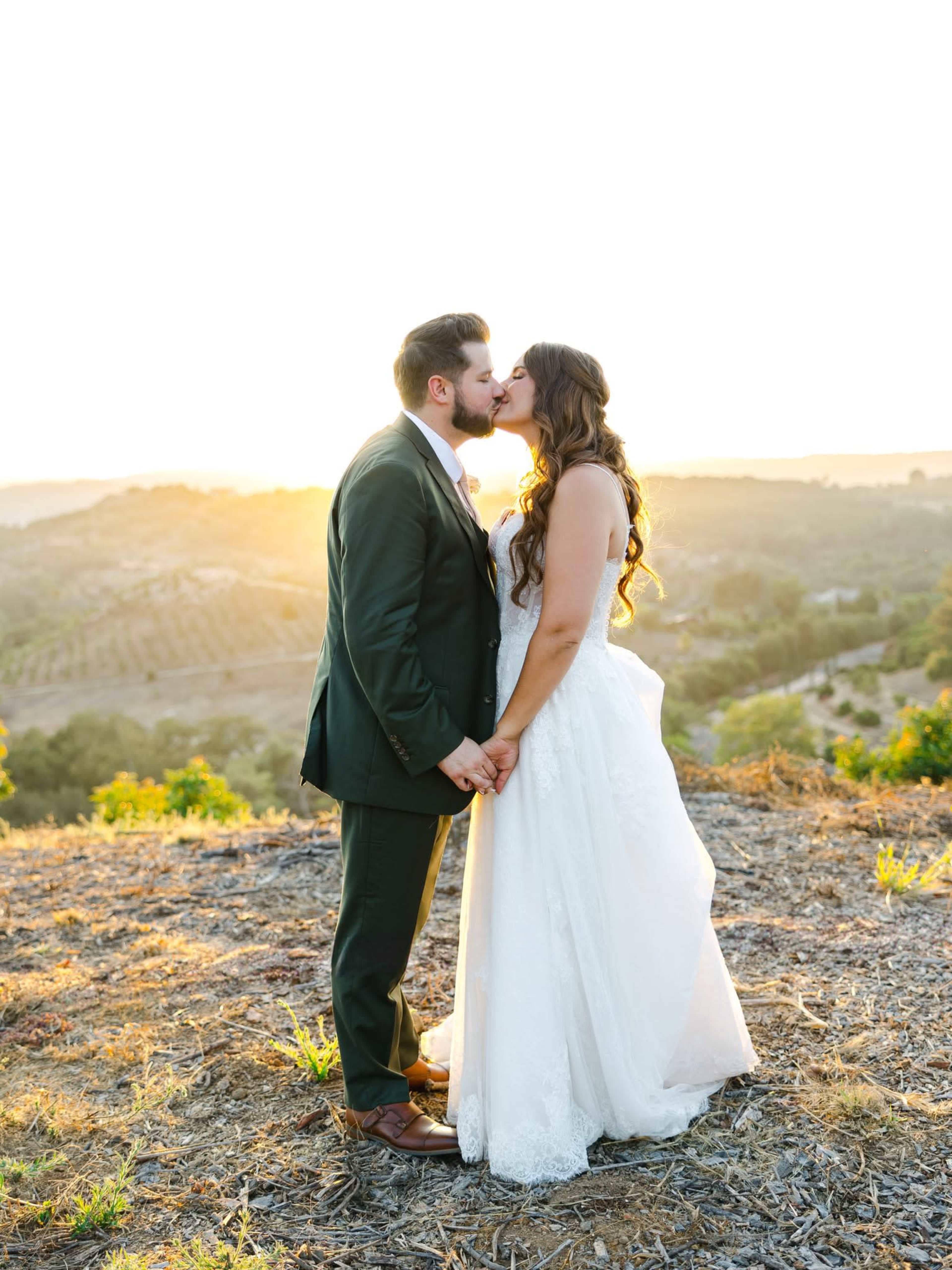 A couple embraces in a kiss during sunset, surrounded by a scenic landscape of hills and vineyards.