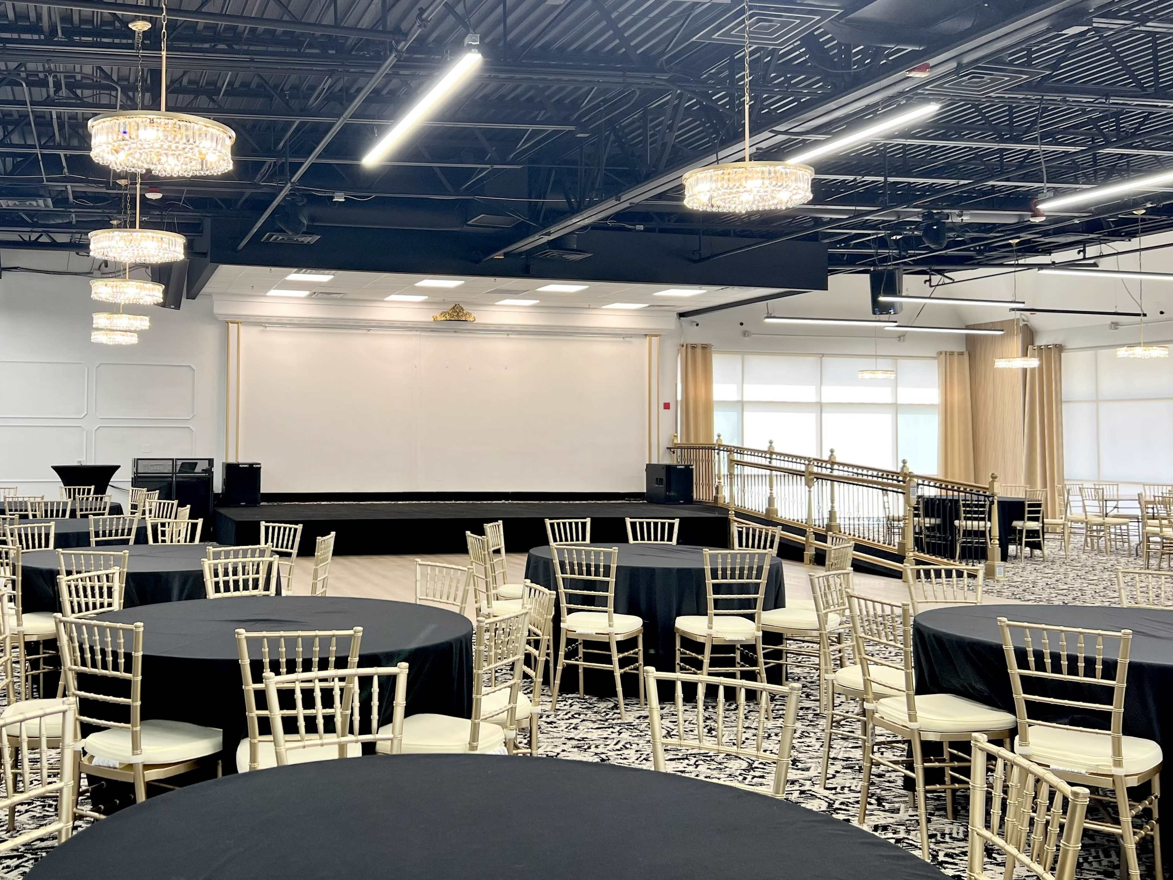 The image shows a banquet hall setup with round tables covered in black tablecloths and gold chairs arranged in front of a stage with white walls and chandeliers overhead.