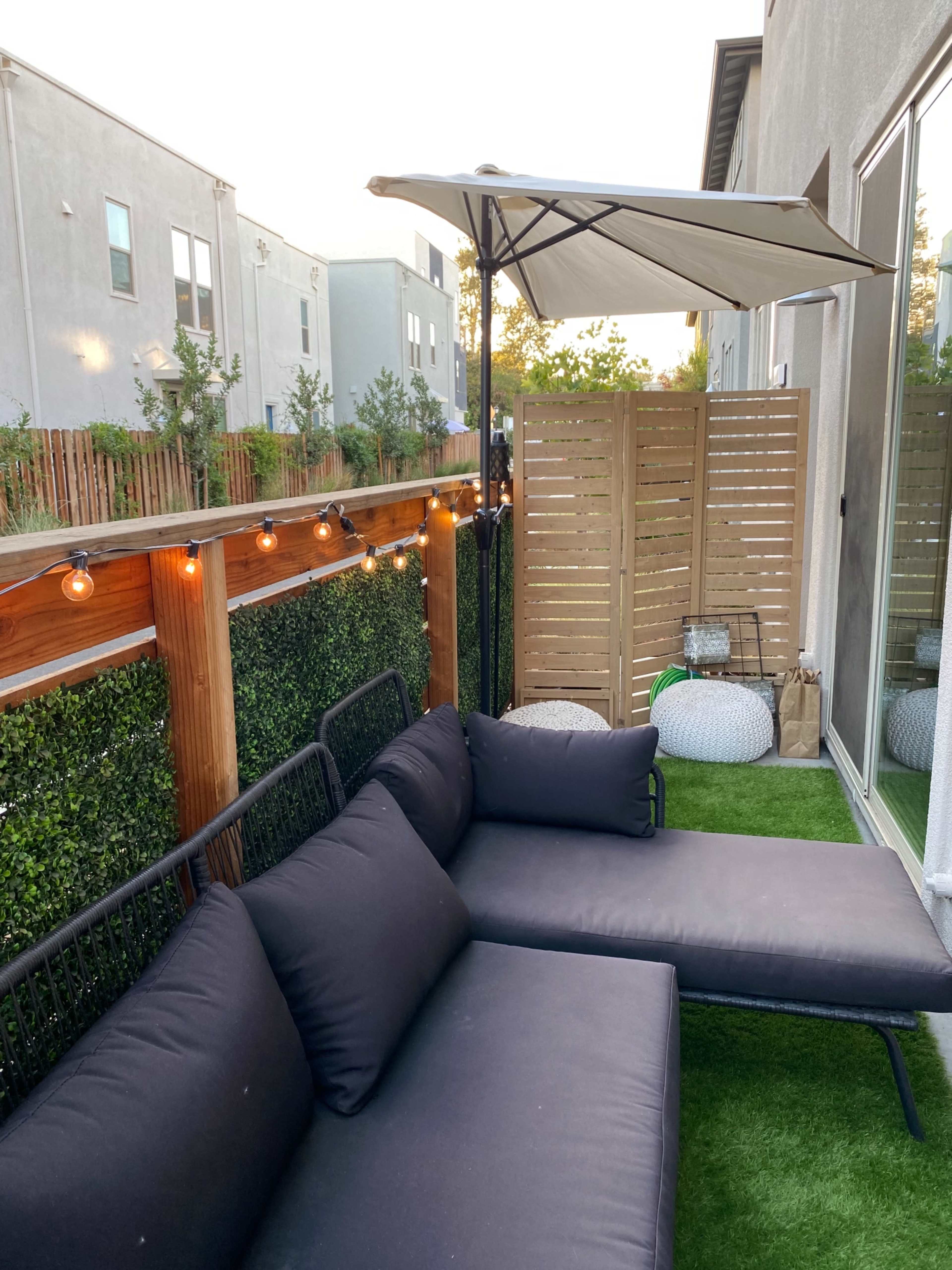 A small outdoor patio area with a black sofa, an umbrella, and wooden fencing, surrounded by grass and decorative lighting.