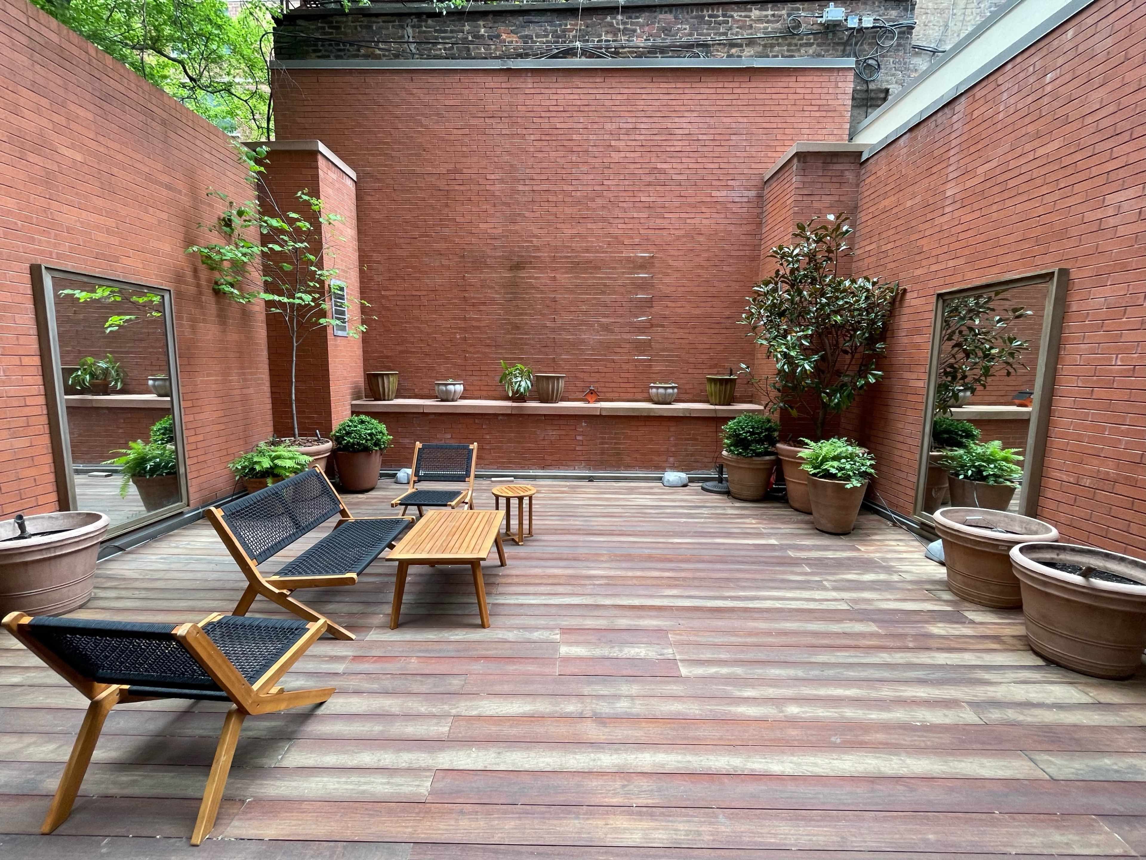 A small outdoor patio with wooden flooring, potted plants, and two seating areas flanked by mirrors on the brick walls.
